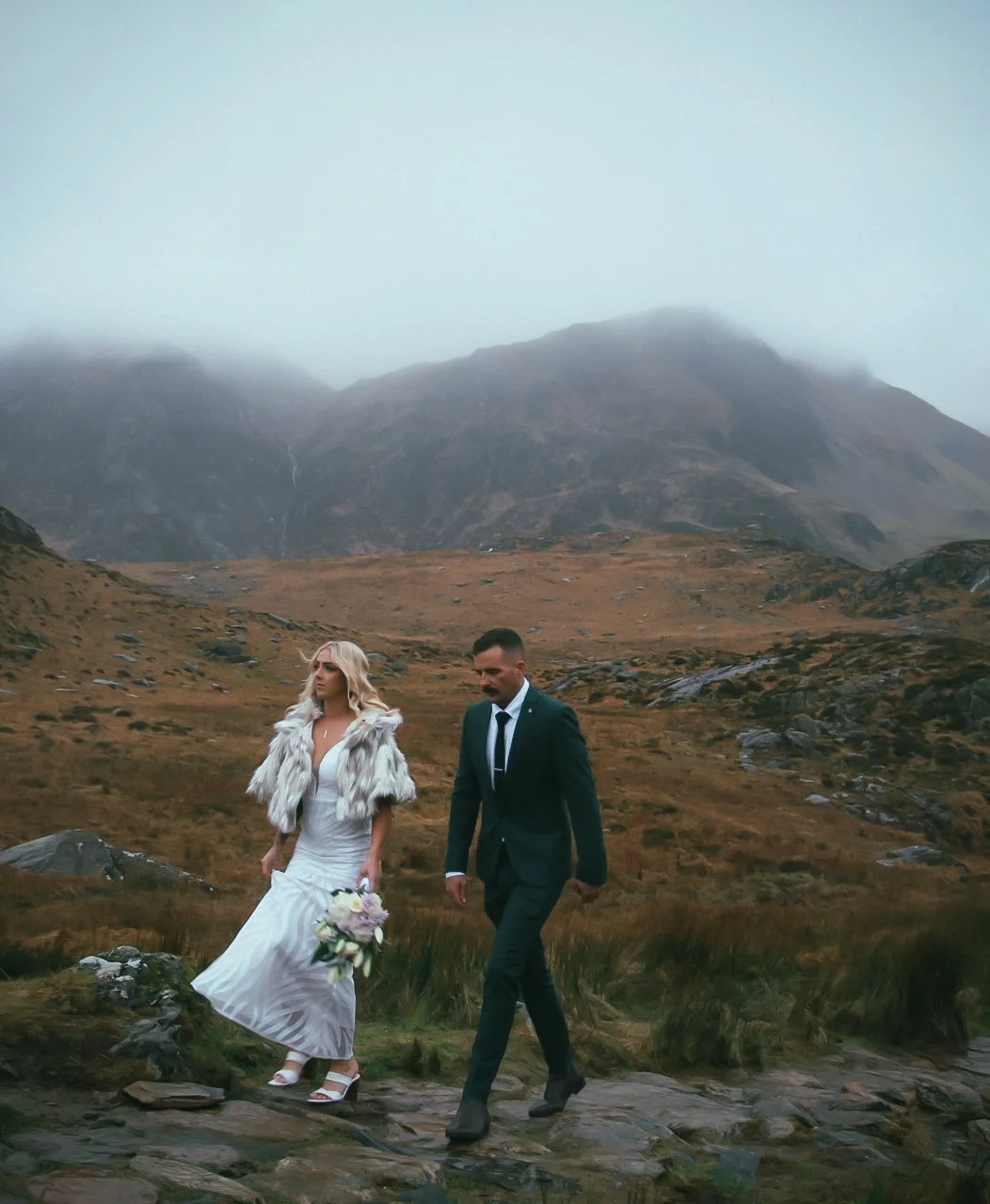A couple walking across rugged moorland beneath misty mountains