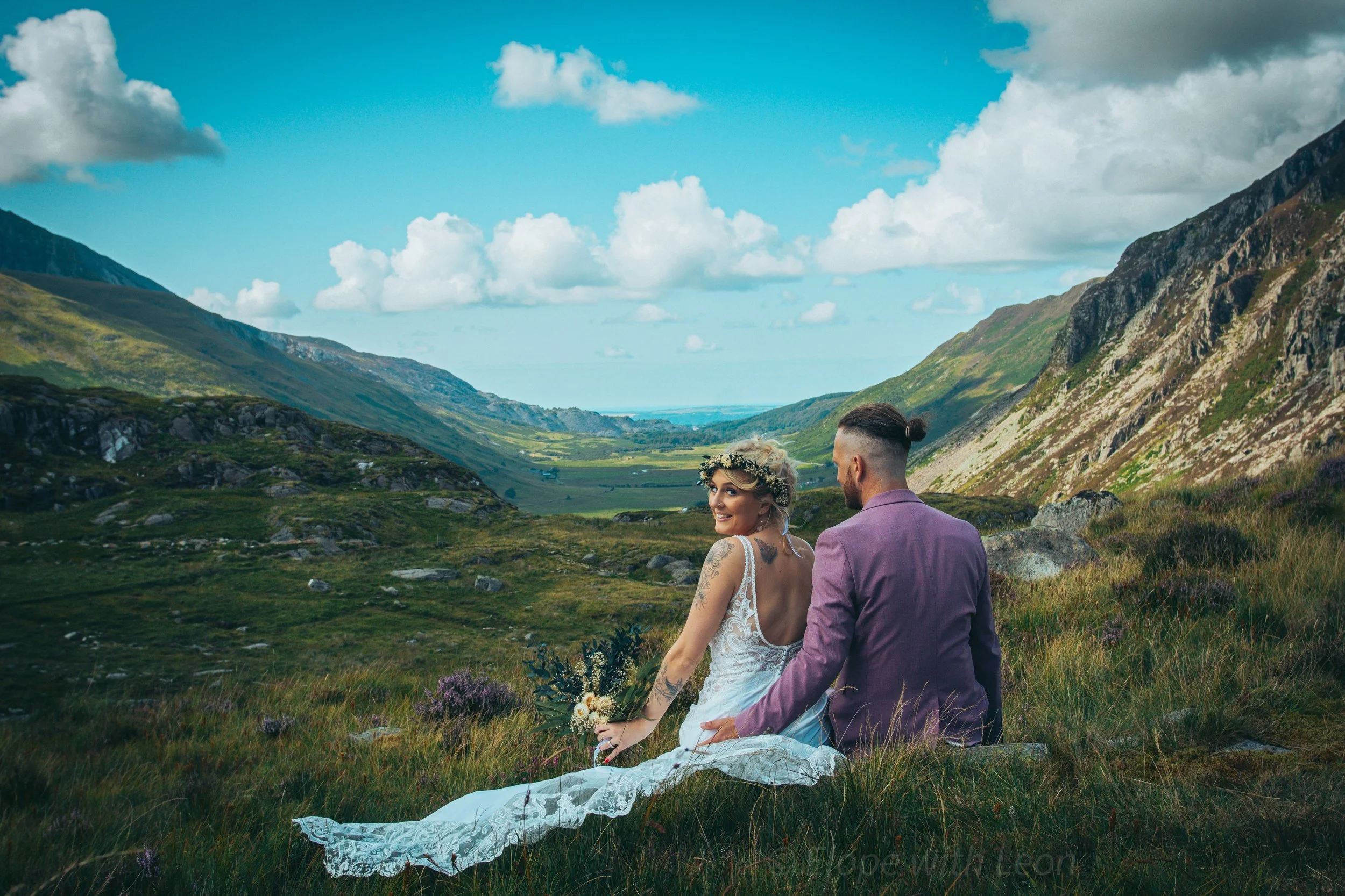 A couple sitting in the grass looking along a green mountain valley