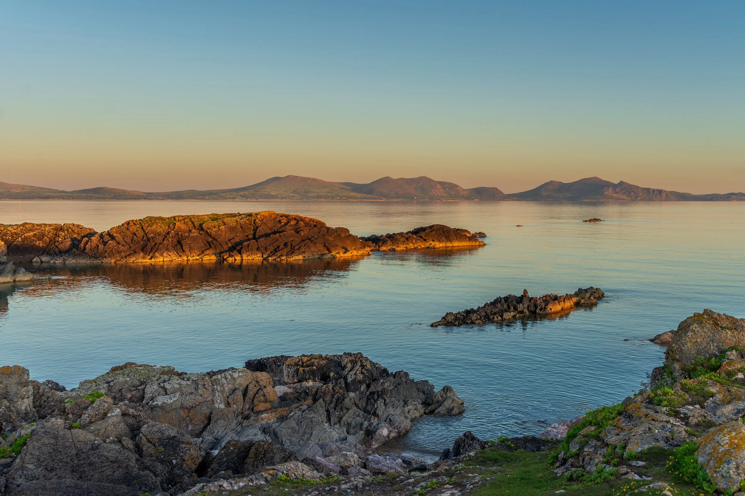 Golden evening light on the rocks and calm water at Llanddwyn Island, with mountains on the horizon