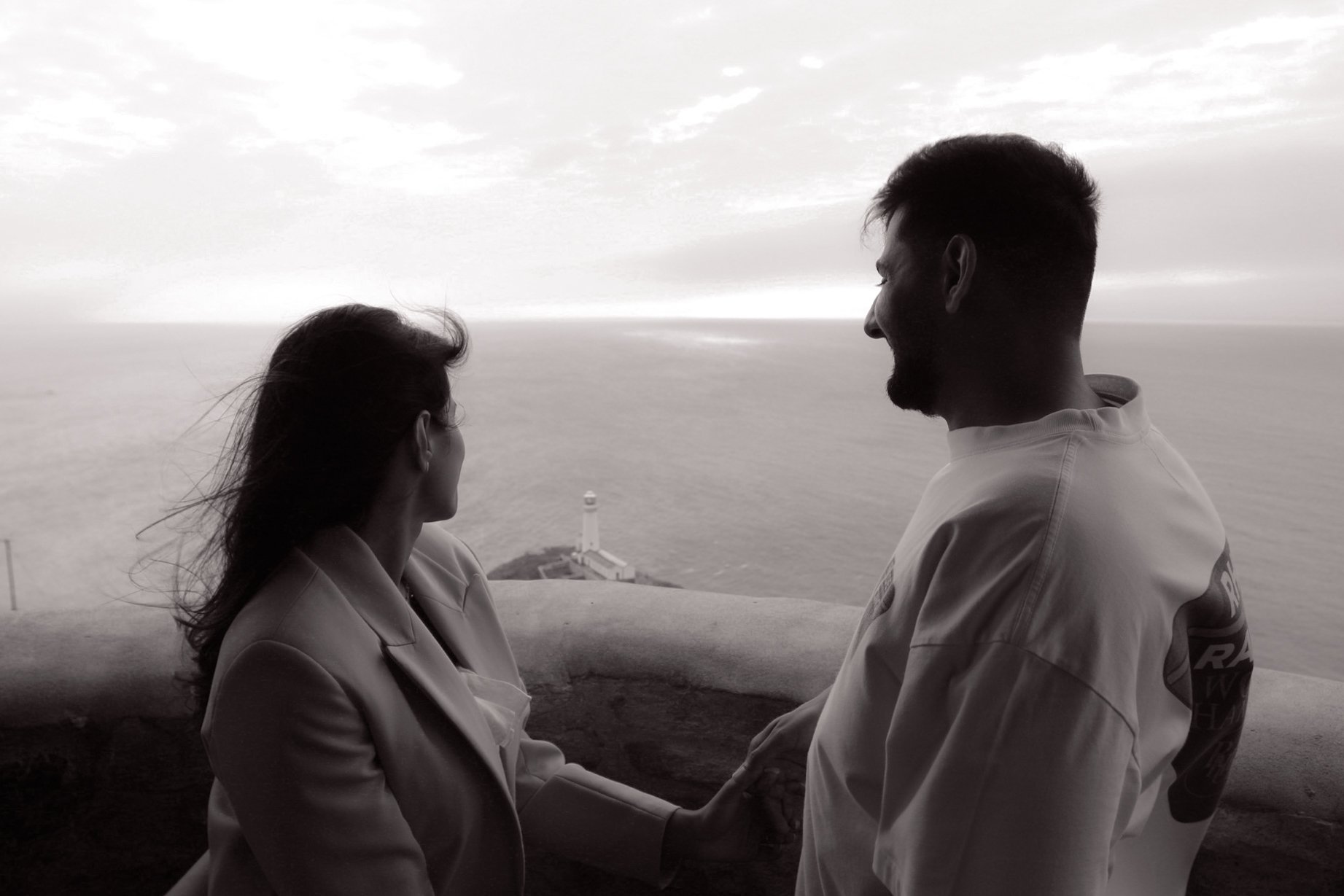 A couple sitting face to face on a clifftop overlooking the sea, in black and white