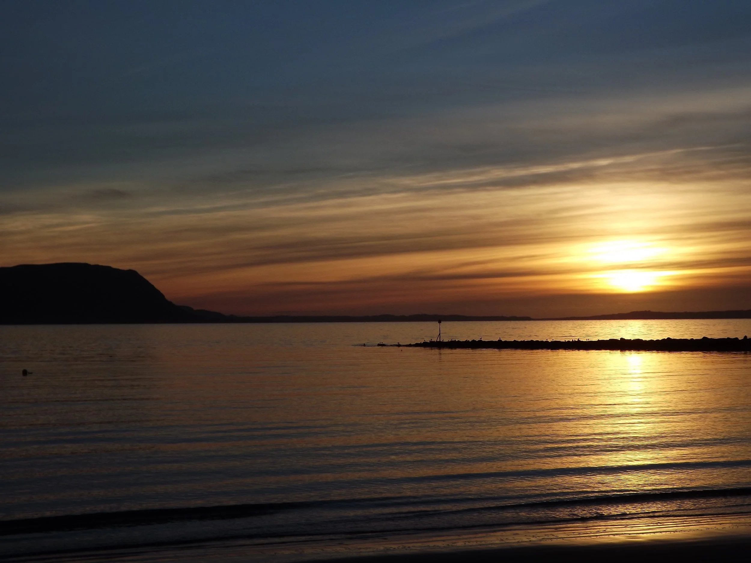 The sun setting in orange and gold over calm water, with the dark outline of land on the horizon, seen from the West Shore