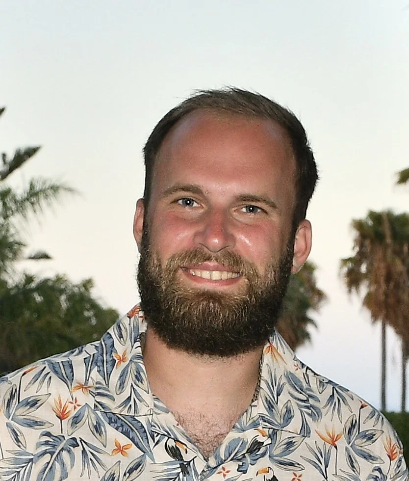 Portrait of Leon Bagnall smiling, with palm trees behind him