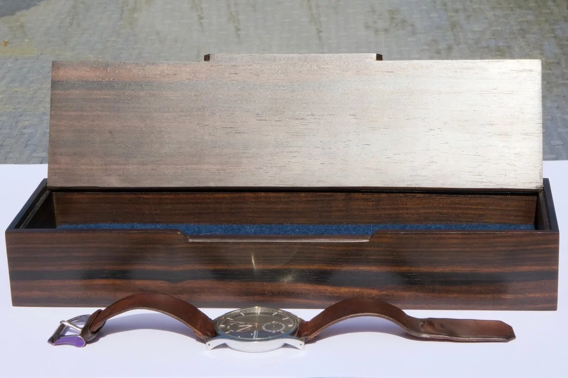 A wooden box with the lid open, placed on a white surface, with a wristwatch with a leather strap laying in front of it.