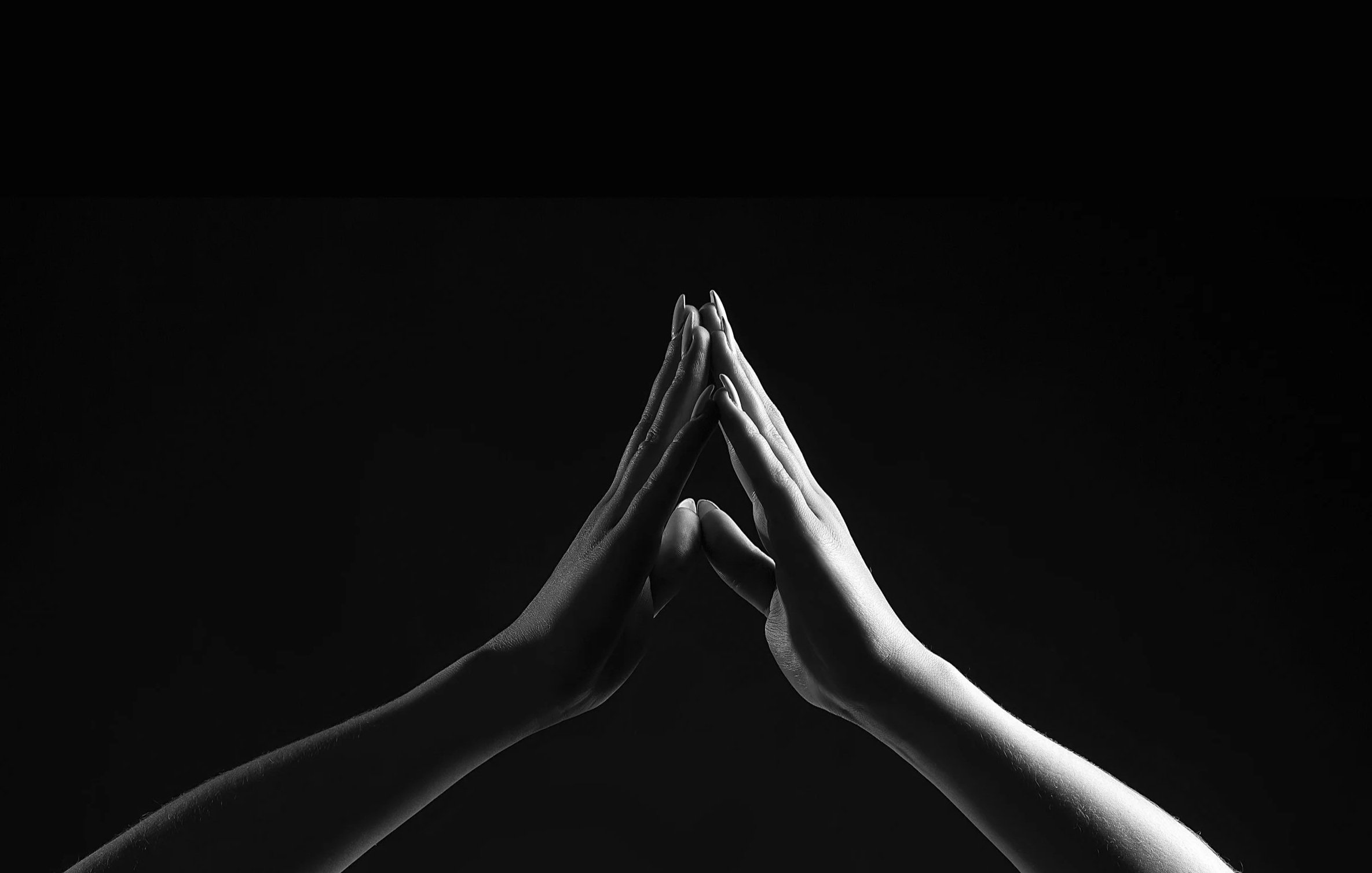 Black and white photograph of two hands pressed together with fingers pointing upward, forming a prayer or greeting gesture against a dark background.