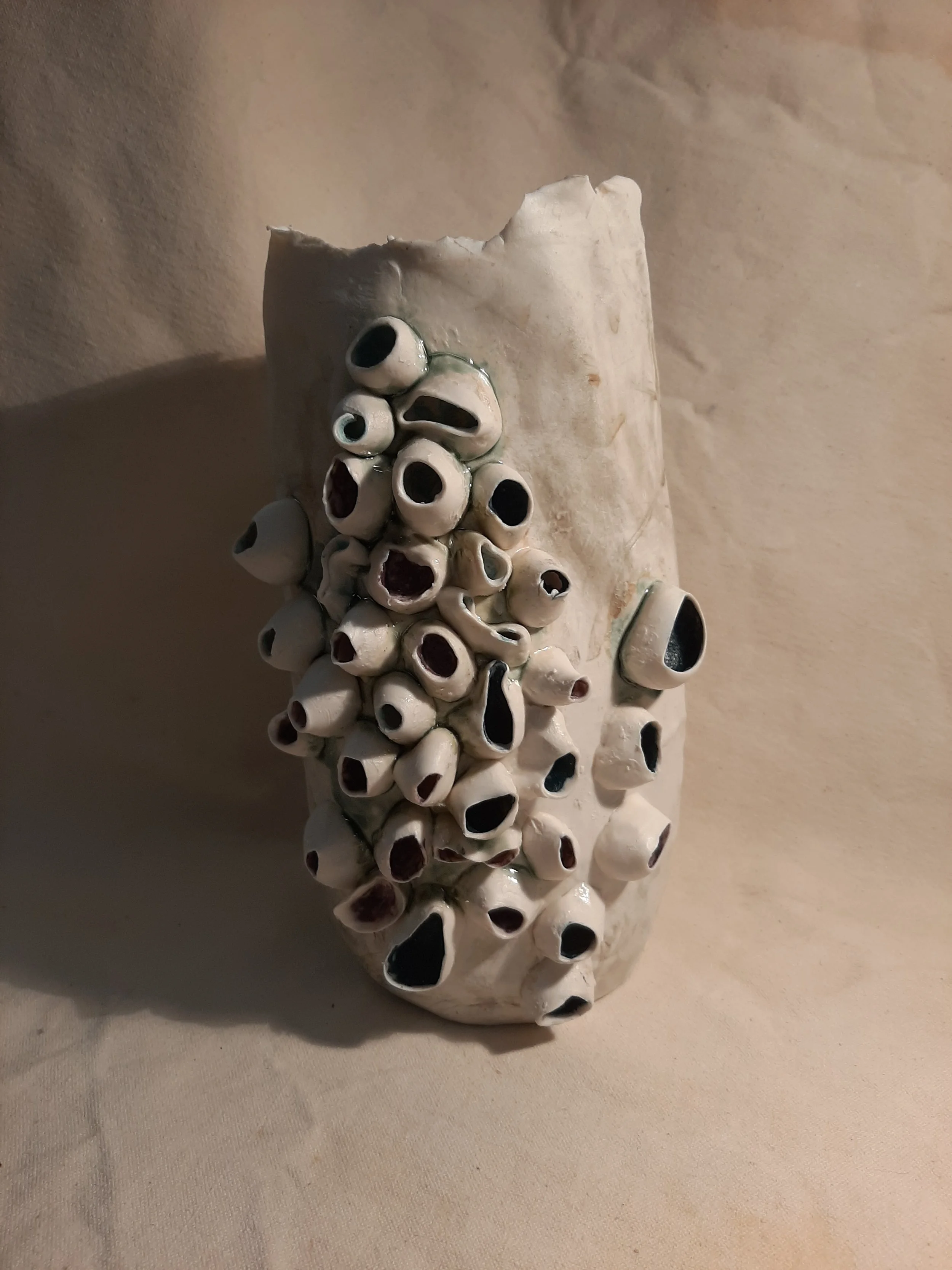 A white ceramic sculpture resembling a cluster of tubular shapes with hollow openings, mounted on a beige fabric surface.