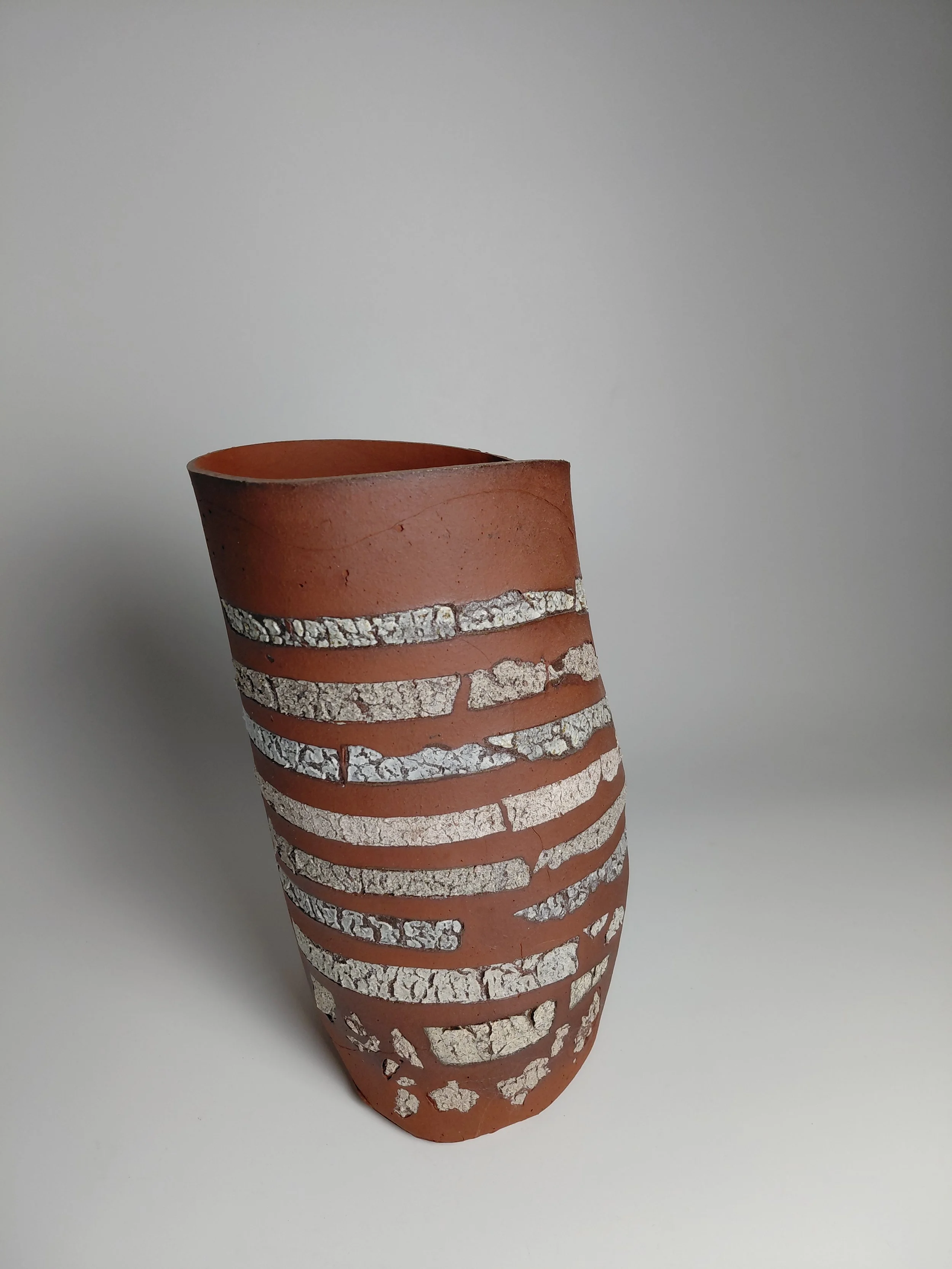 A tall, rugged, orange clay vase with white and gray textured horizontal stripes on a light gray background.