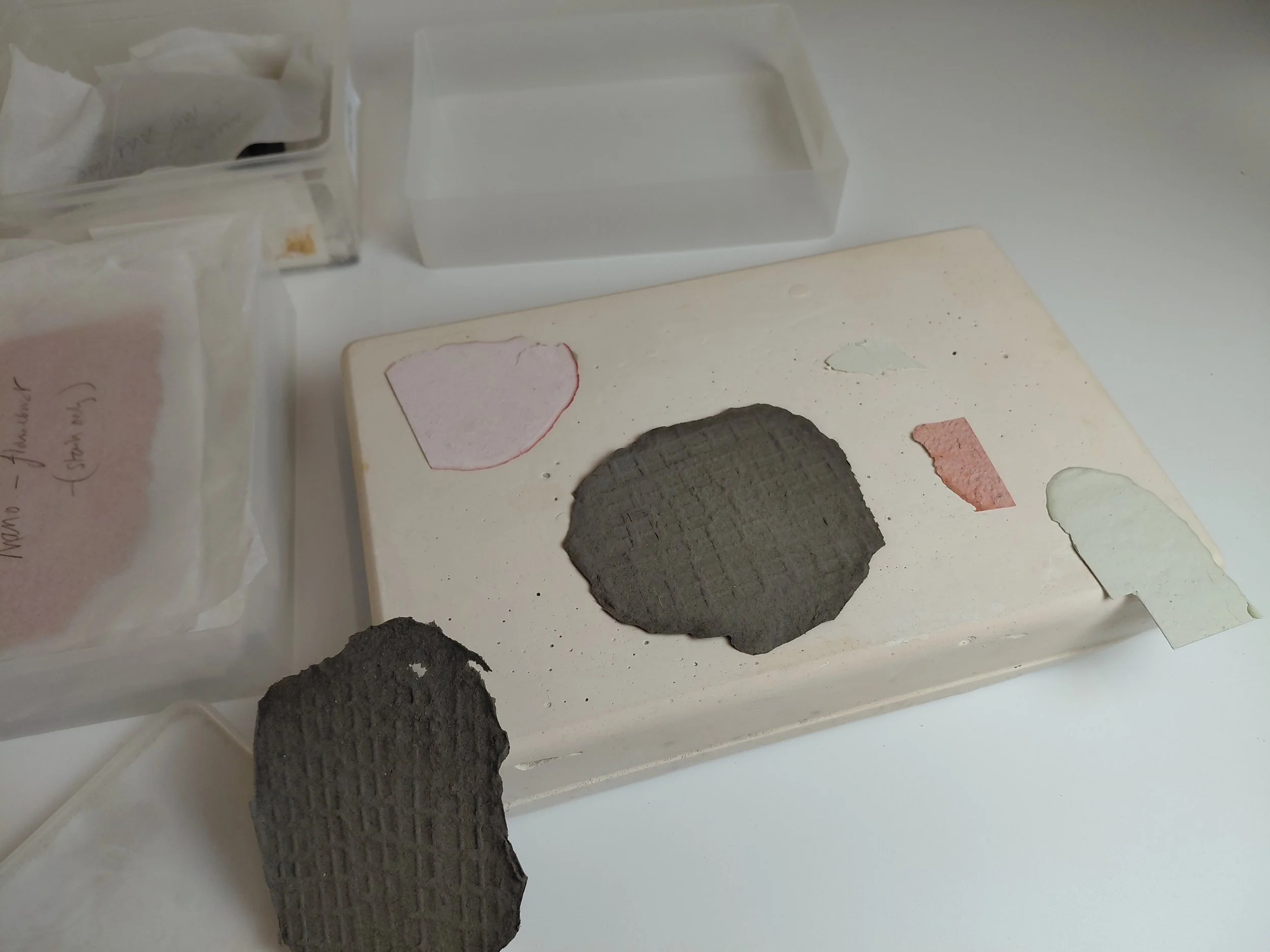 A collection of paint chips and samples on a white surface, including a black textured circle, beige, pink, white, and coral-colored pieces, with packaging visible on the left.