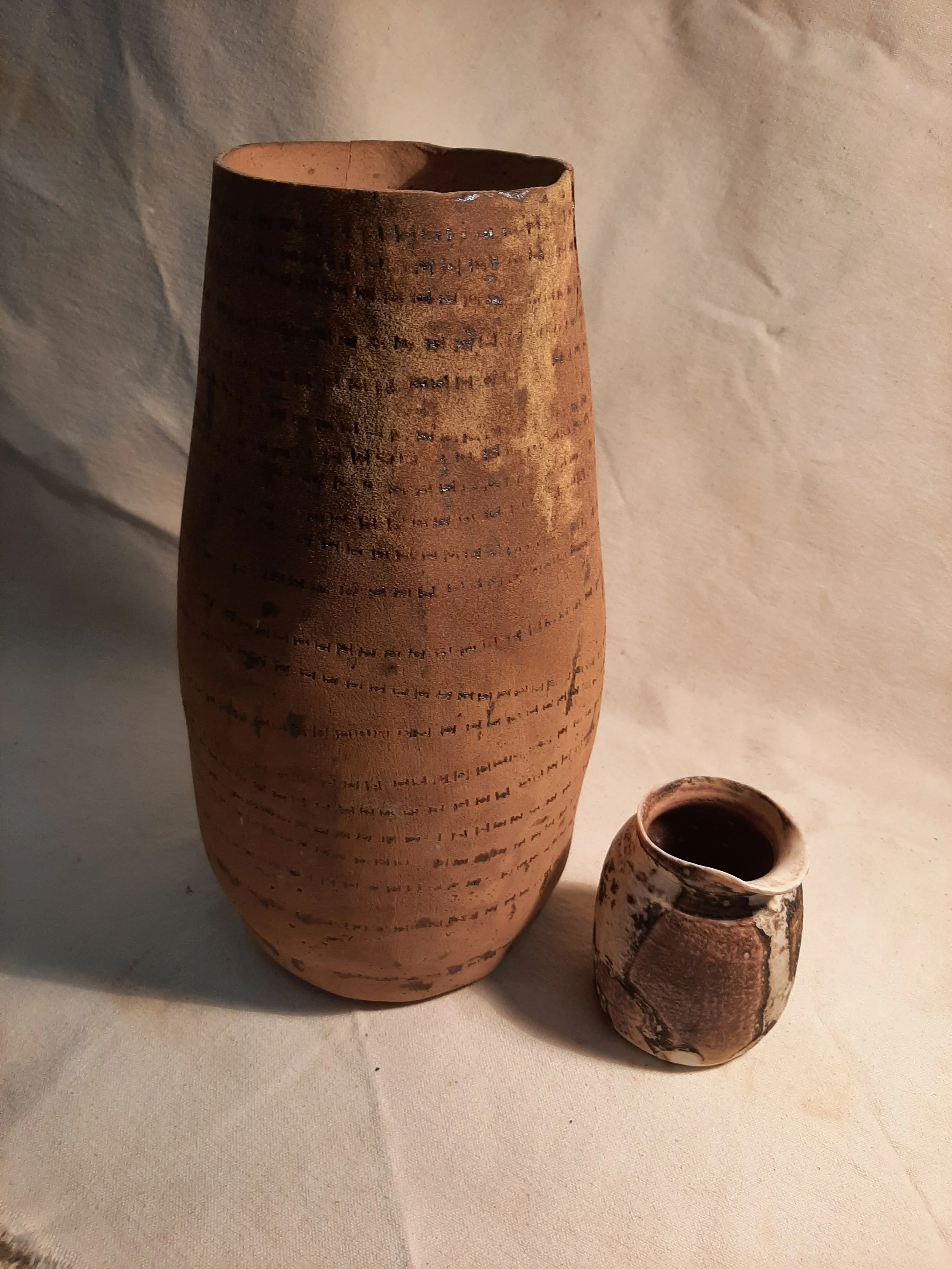Two ancient pottery jars with inscriptions, one tall and narrow and the other small and rounded, placed on a beige surface against a plain wall.
