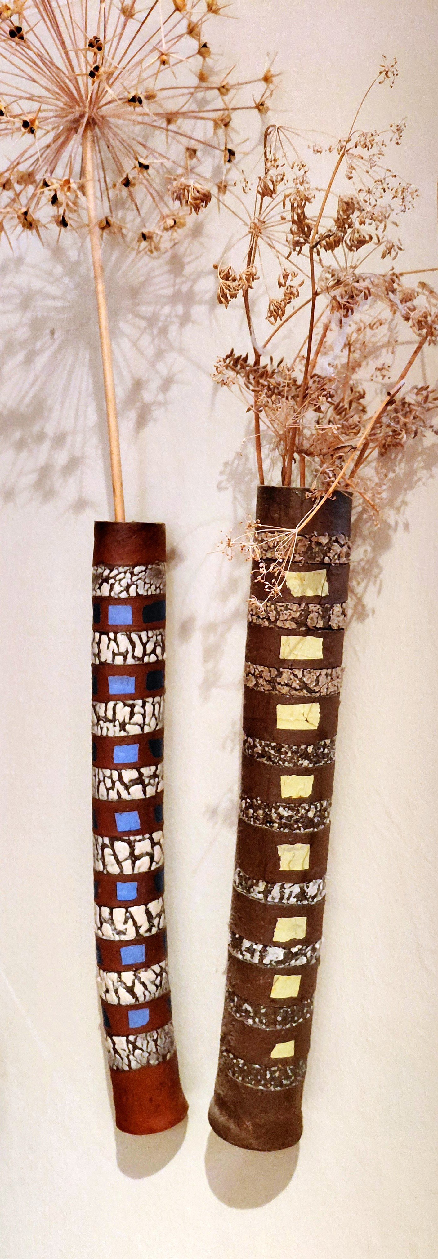 Two tall decorative vases with dried plants. The vases have geometric designs and patterns, with colors including brown, black, white, blue, and yellow.