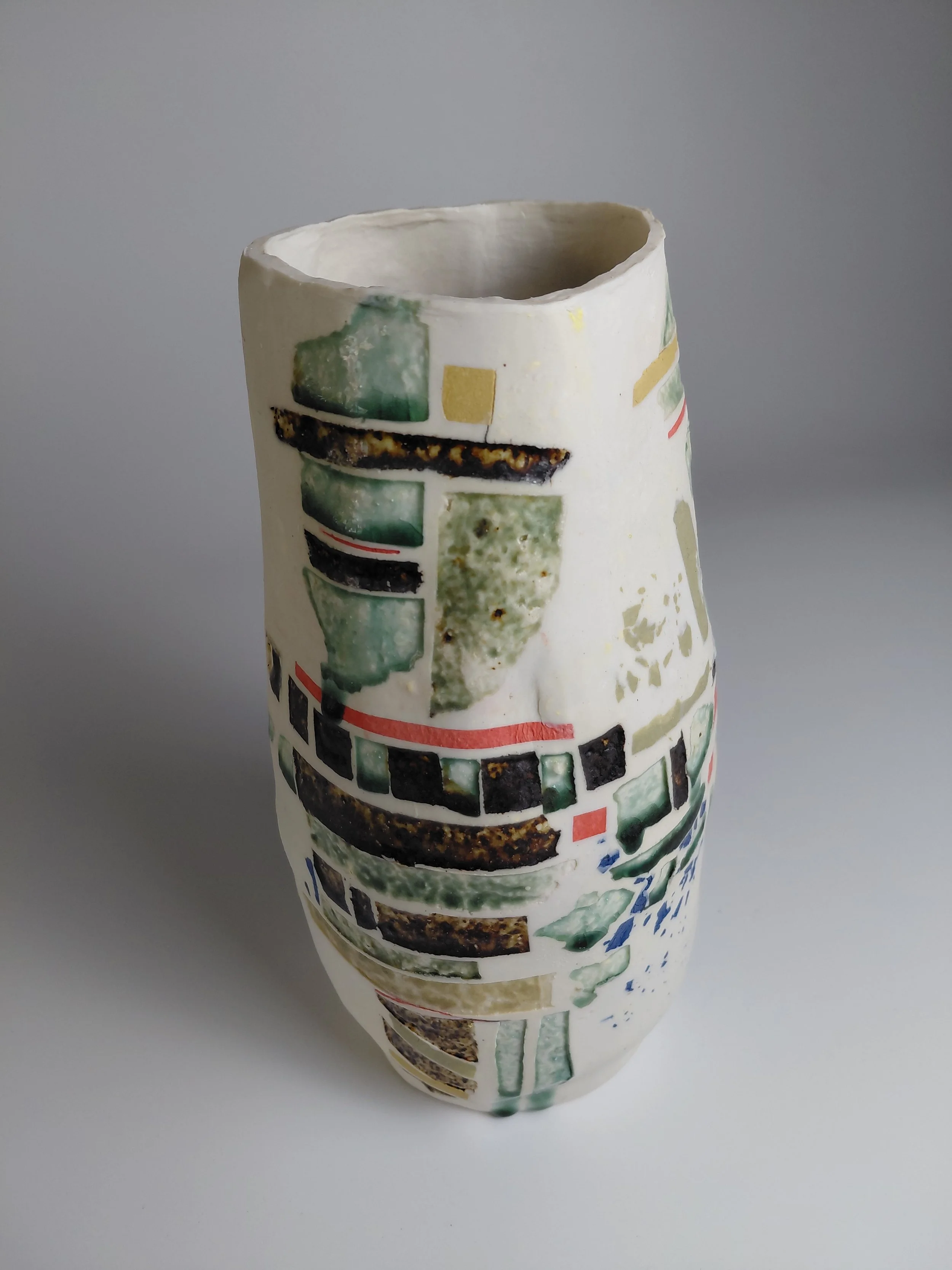 A tall ceramic vase with an irregular top edge, decorated with abstract, colorful rectangular patterns in green, yellow, red, black, and white, on a light gray background.