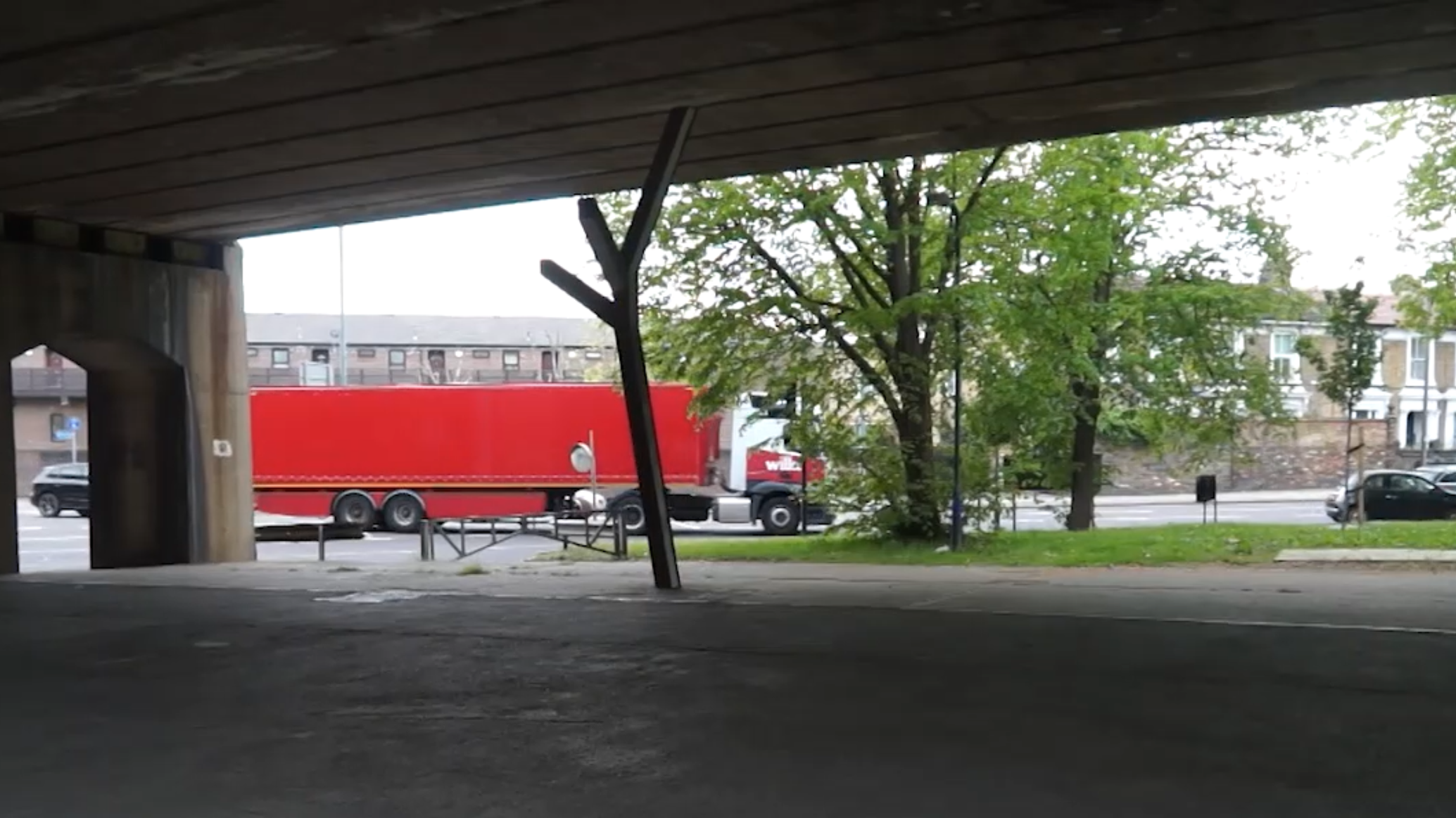 View from under a bridge showing a street with a red truck, trees, and residential buildings in the background.