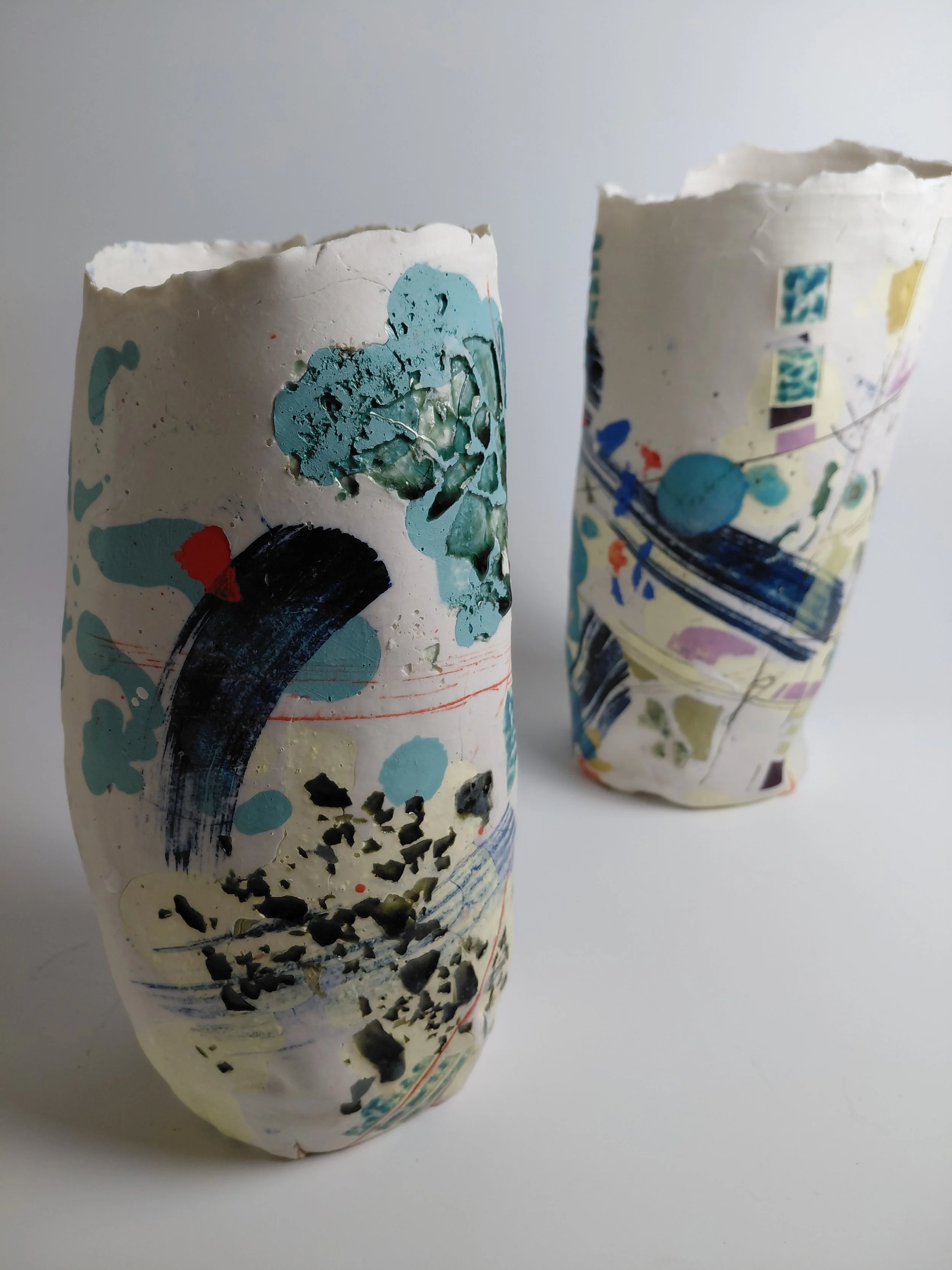 Two paper cups with colorful abstract painted designs, one with more visible black and red splashes, placed on a white surface against a neutral background.