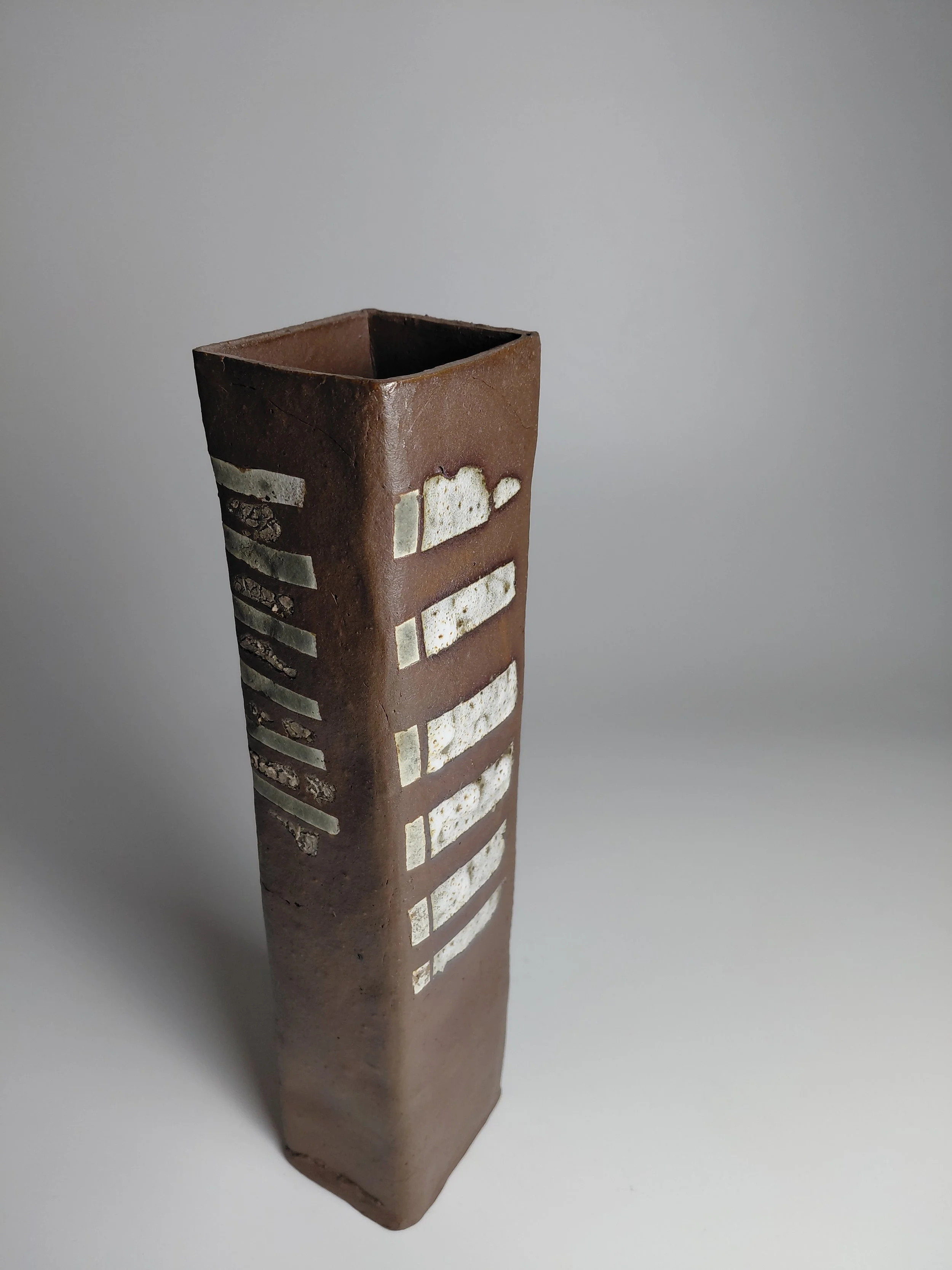 A tall, rectangular ceramic vase with a brown glaze and decorative white accents, placed against a plain gray background.