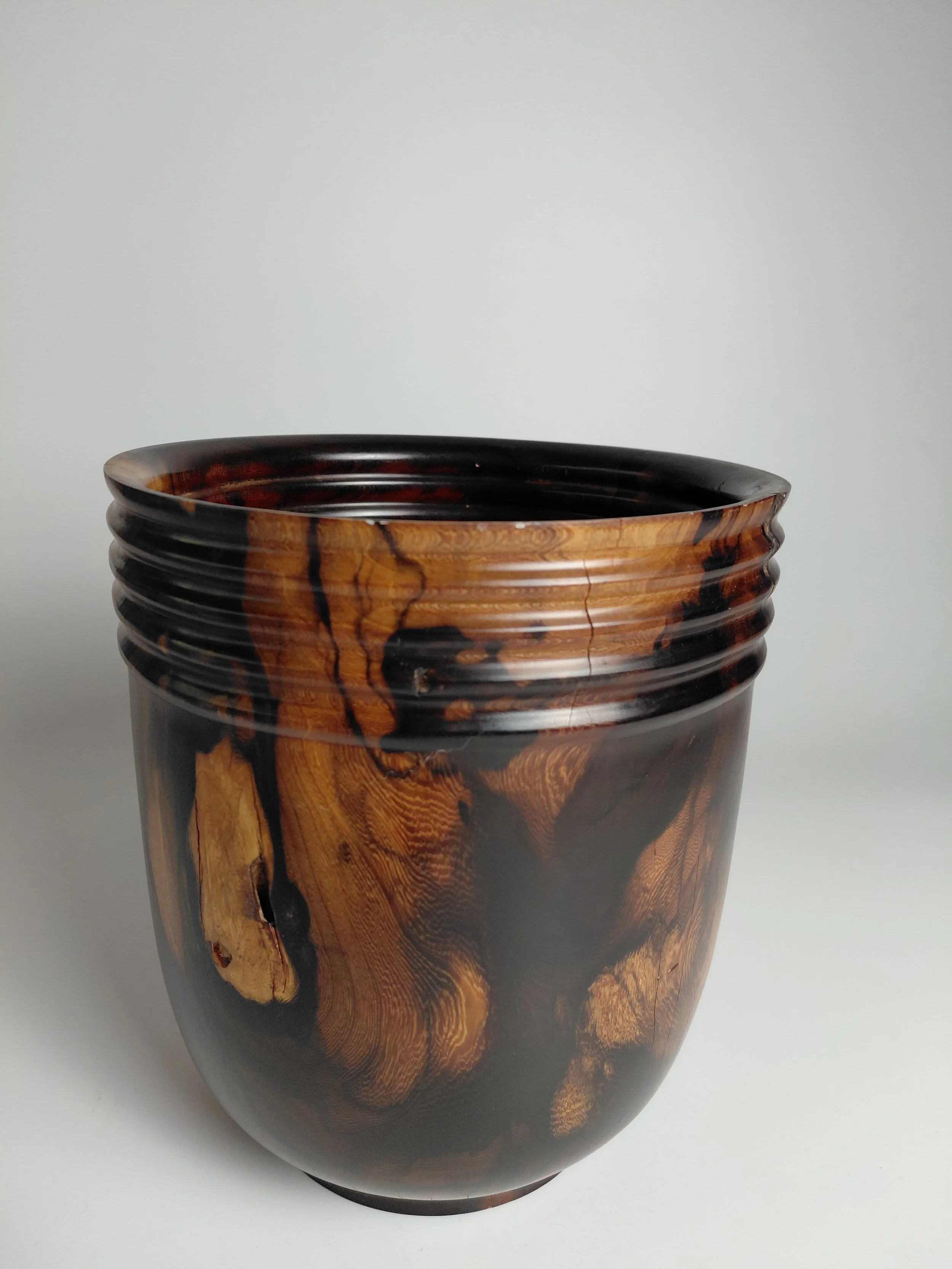 A tall, cylindrical wooden vase with a glossy finish showcases a dark, rich wood grain pattern with swirling natural designs, set against a plain light background.