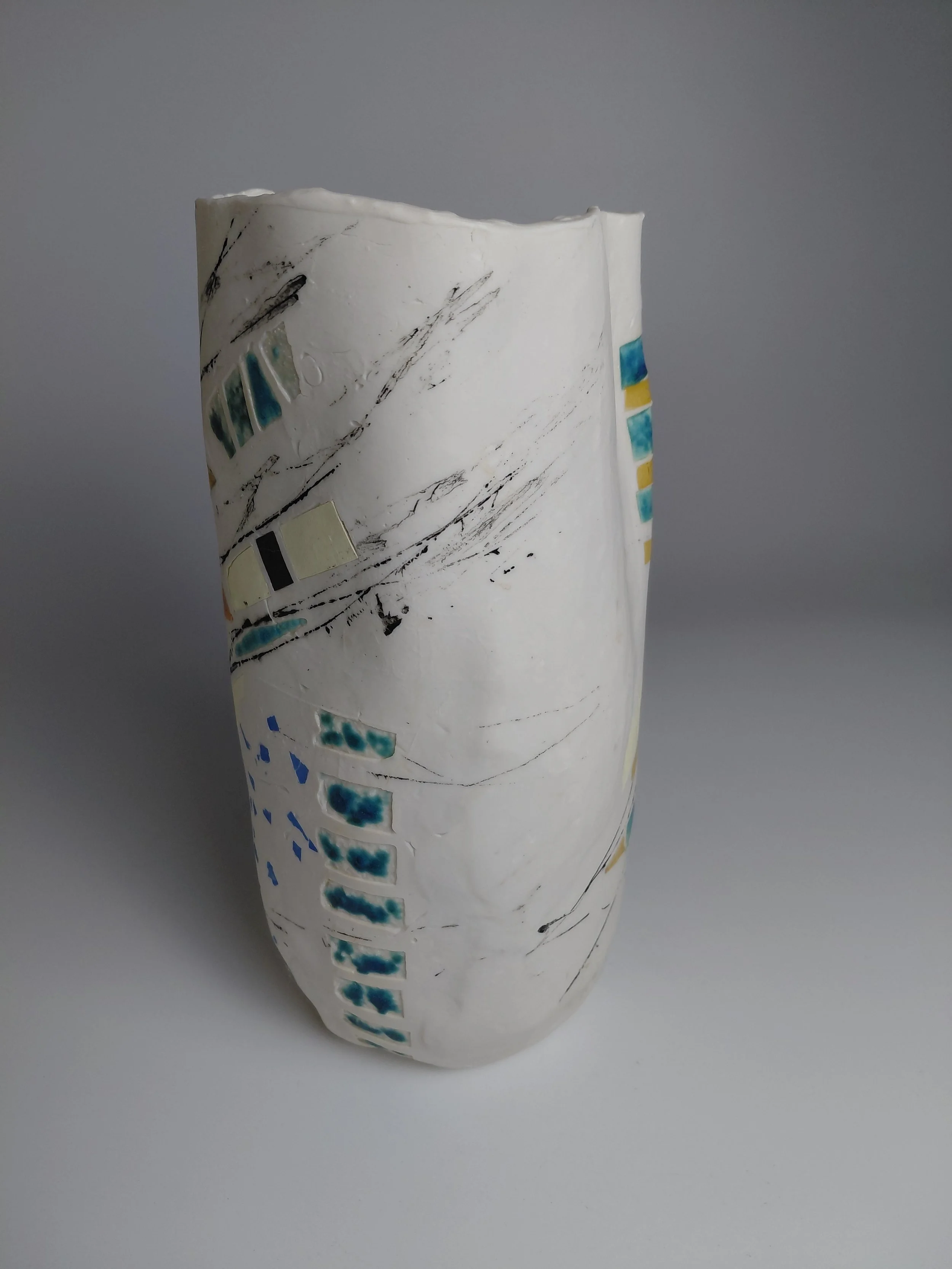Close-up of a rolled-up white sheet of paper with abstract black sketch lines and small rectangular blotches of blue, yellow, black, and beige markings, against a plain gray background.