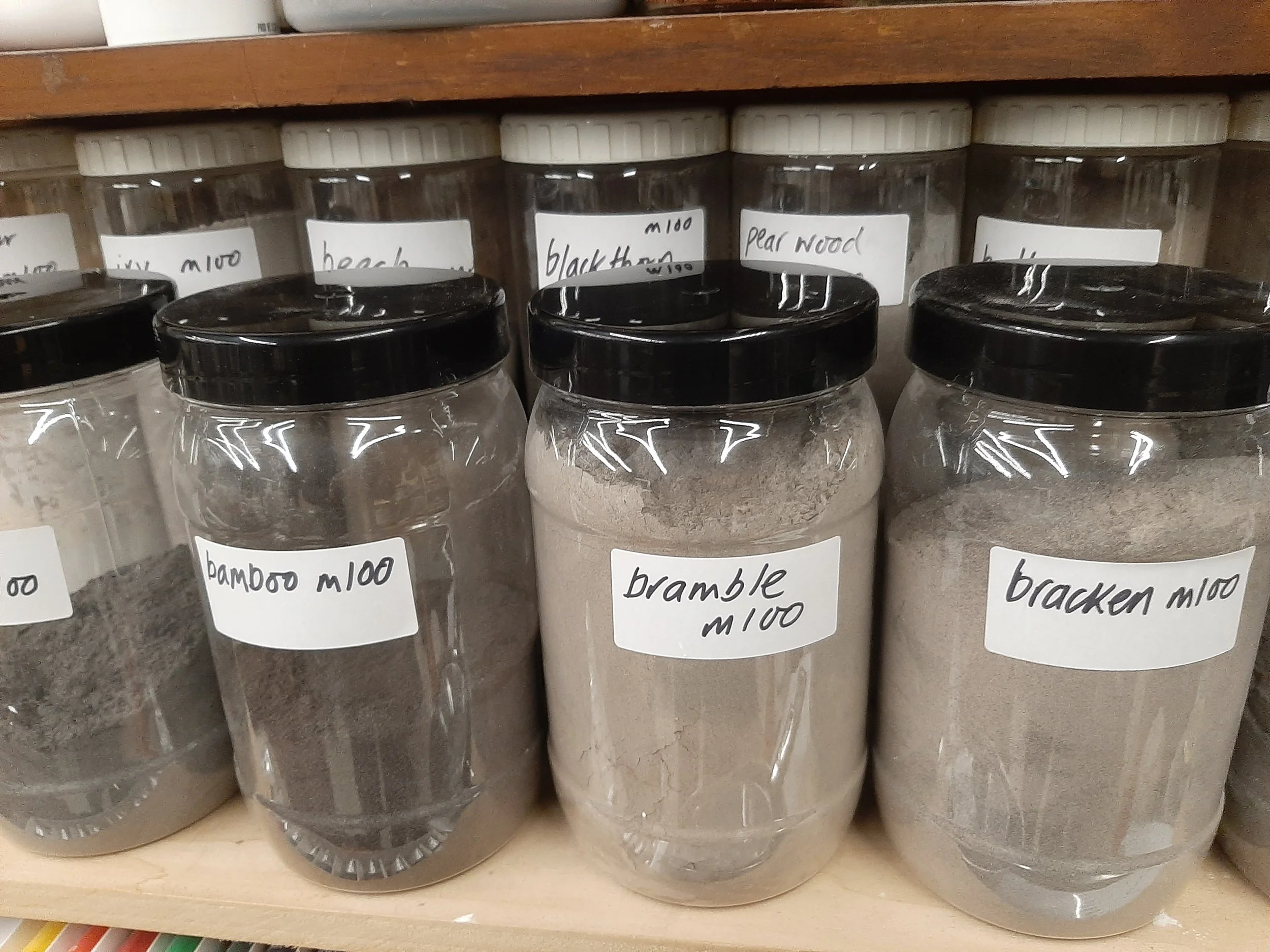 Several clear jars with black lids containing plant ashes, labeled with handwritten signs indicating different types such as bamboo, bramble, blackthorn, and pear wood, on a wooden shelf.