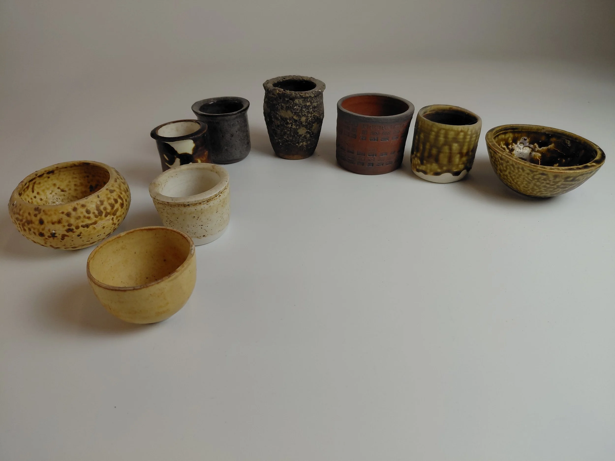 A collection of small, irregularly shaped ceramic cups and bowls in various colors and textures arranged on a white surface against a plain background.