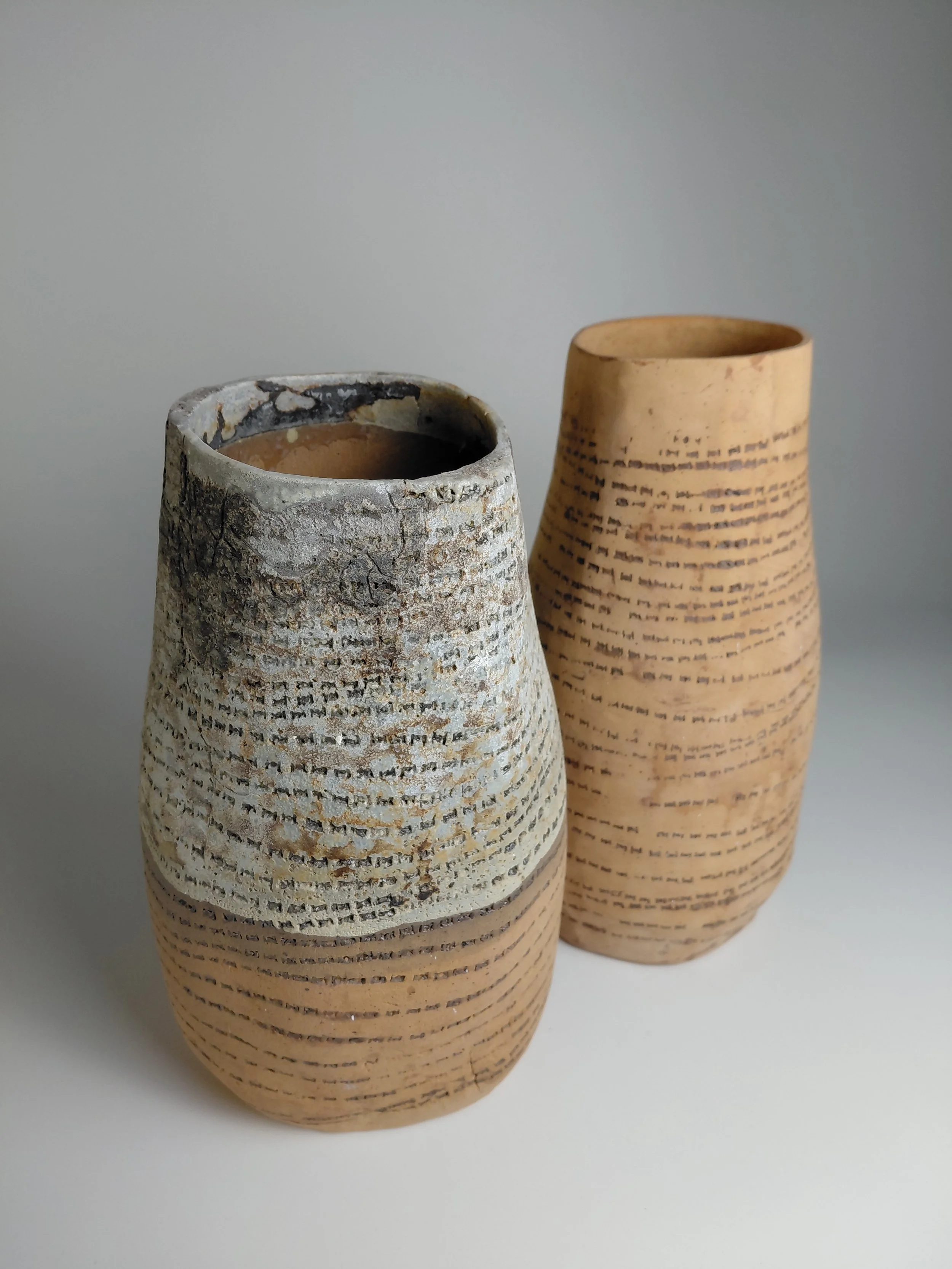 Two ceramic vases with textured and patterned surfaces, one in grayish tones and the other in light tan, placed against a plain background.