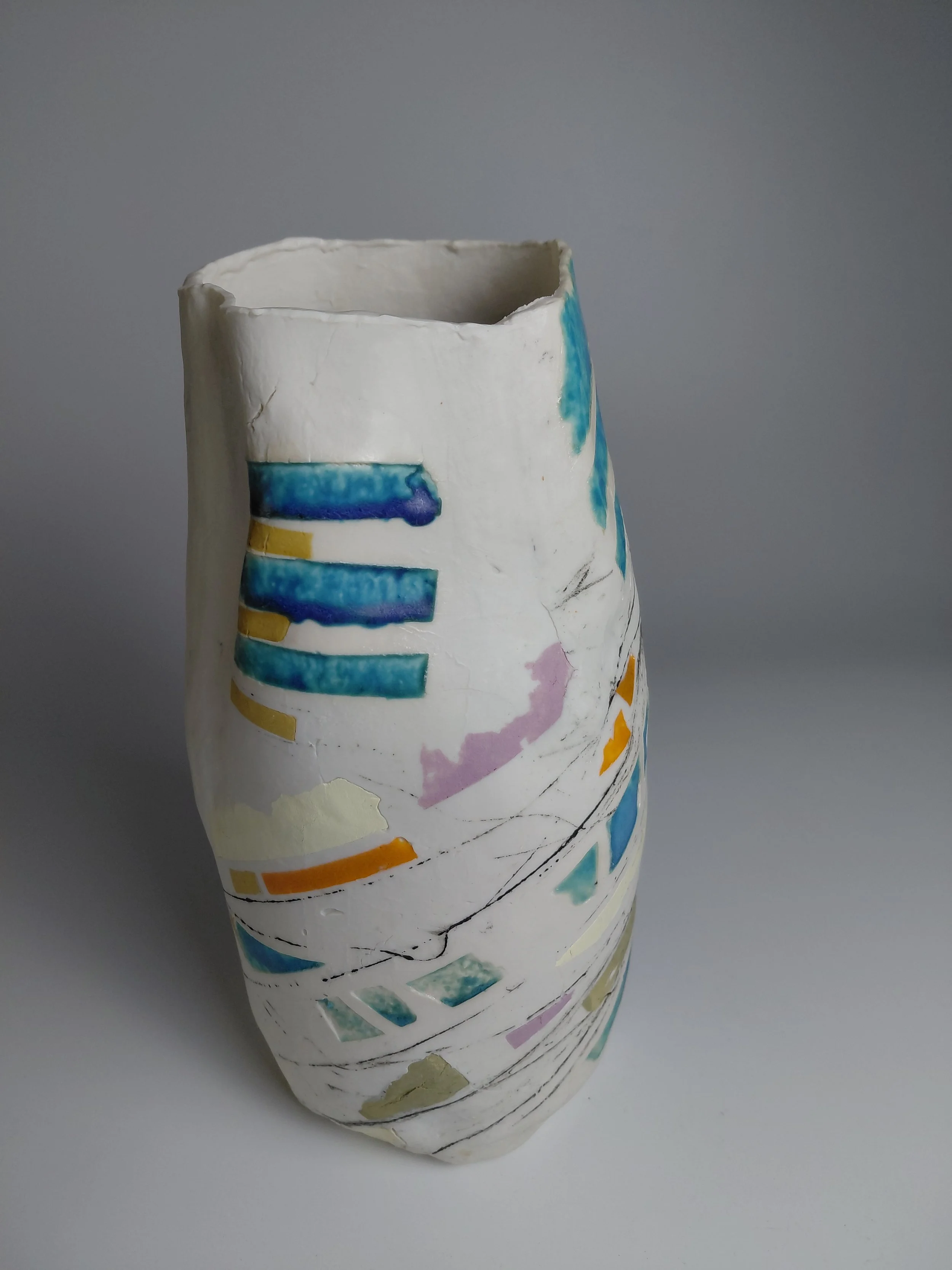 Ceramic vase with hand-painted colorful geometric designs on a white background, positioned against a plain gray backdrop.