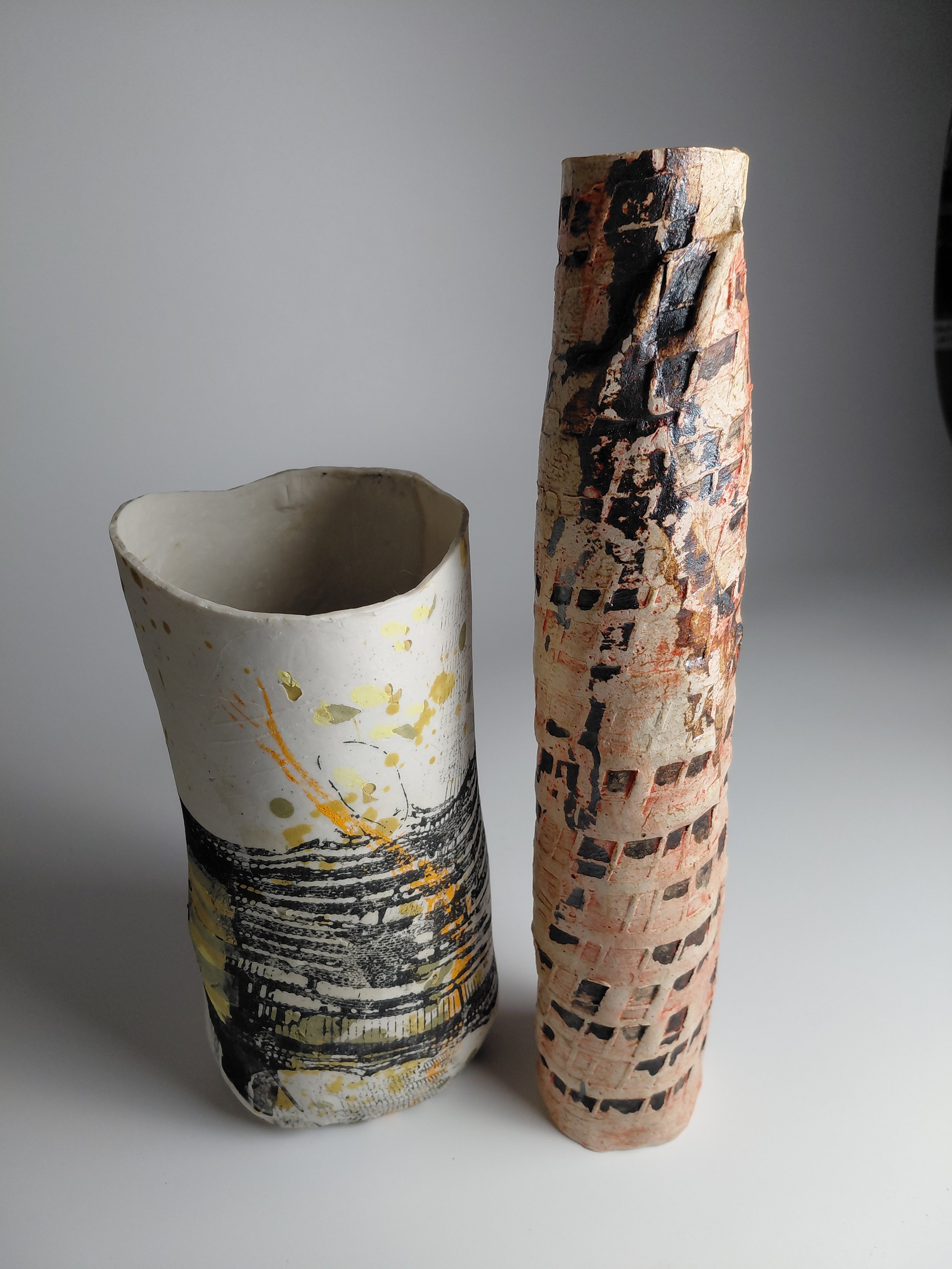 Two ceramic vases with abstract artistic designs, one with black, yellow, and white patterns, and the other with a textured reddish-orange and black pattern, against a plain gray background.