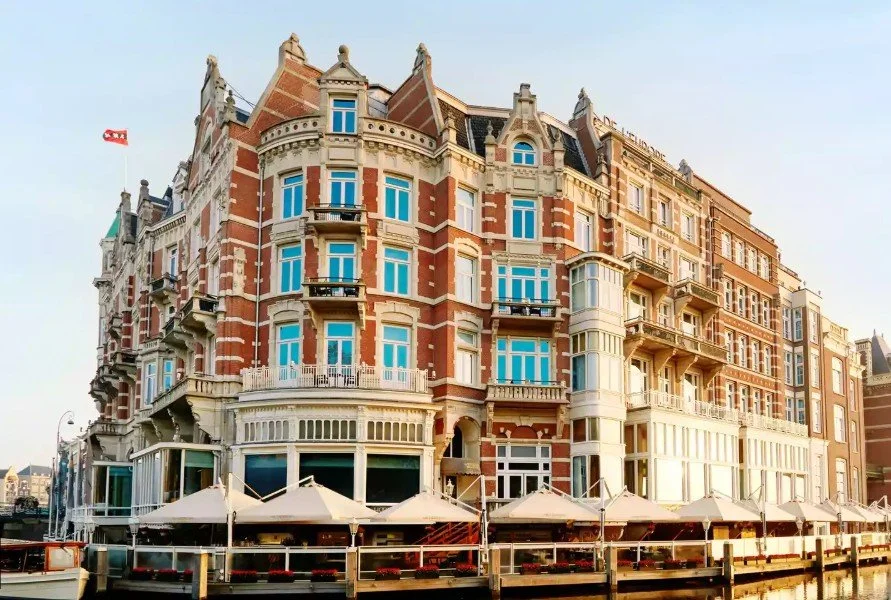 Historic brick building with ornate architectural details, balconies, and large windows, situated along a waterfront with outdoor seating and umbrellas.