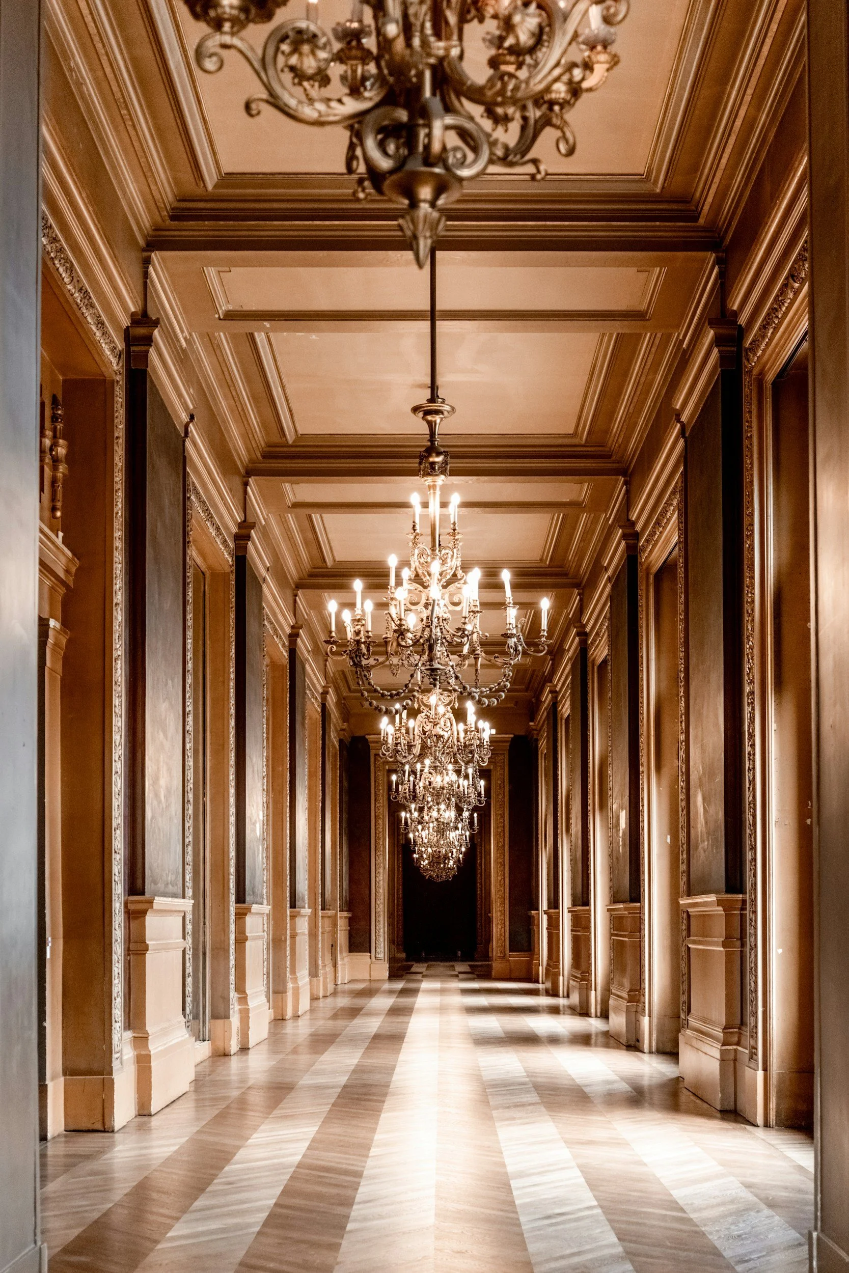 Elegant corridor with ornate chandeliers hanging from a coffered ceiling, gold-toned walls with intricate moldings, and a patterned wooden floor.