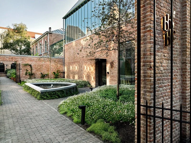 A modern building with a brick exterior and glass upper level, surrounded by well-maintained greenery, including small trees and white flowering plants. A fountain is situated along a paved walkway, with a black metal fence seen on the right side.