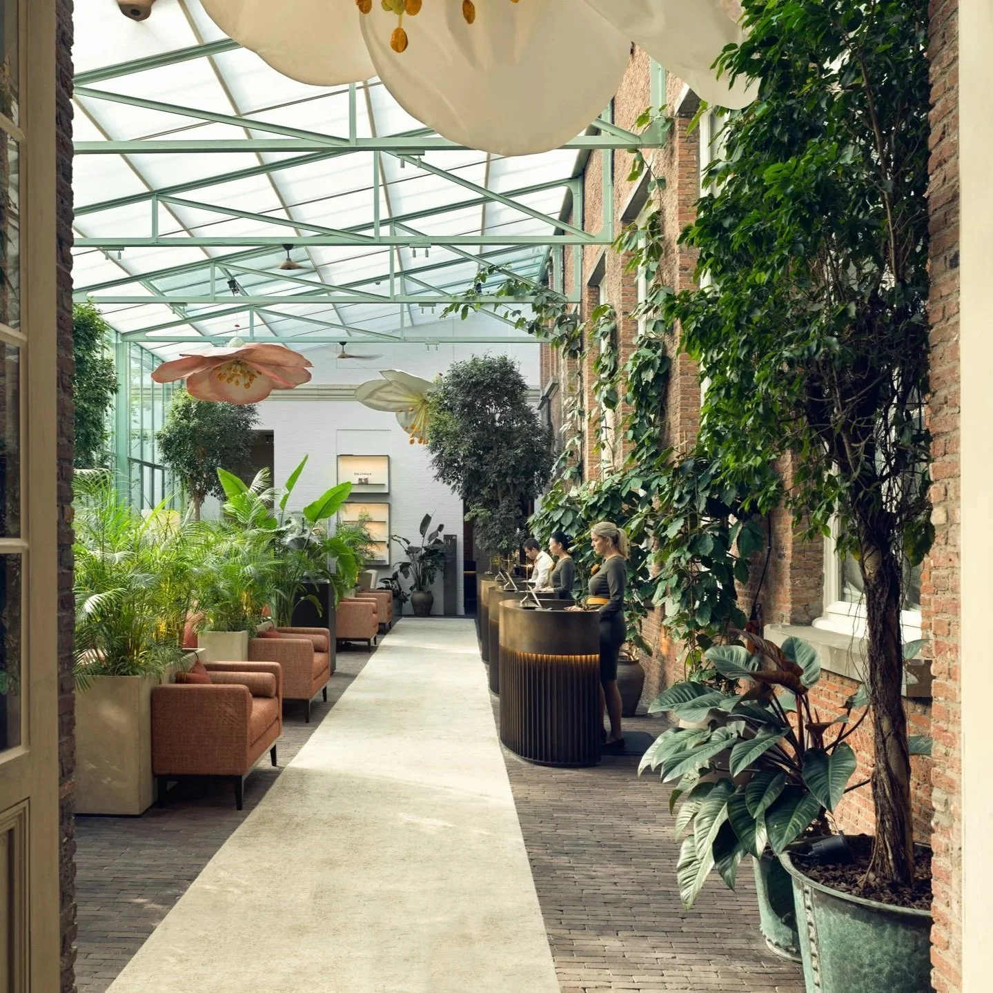 Indoor botanical lounge with large plants, pink and white floral hanging decorations, and a reception desk with staff using computers.