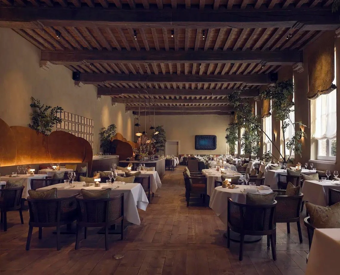 Elegant restaurant interior with decorated tables, white tablecloths, glasses, candles, large windows with curtains, wooden ceiling, and potted plants.