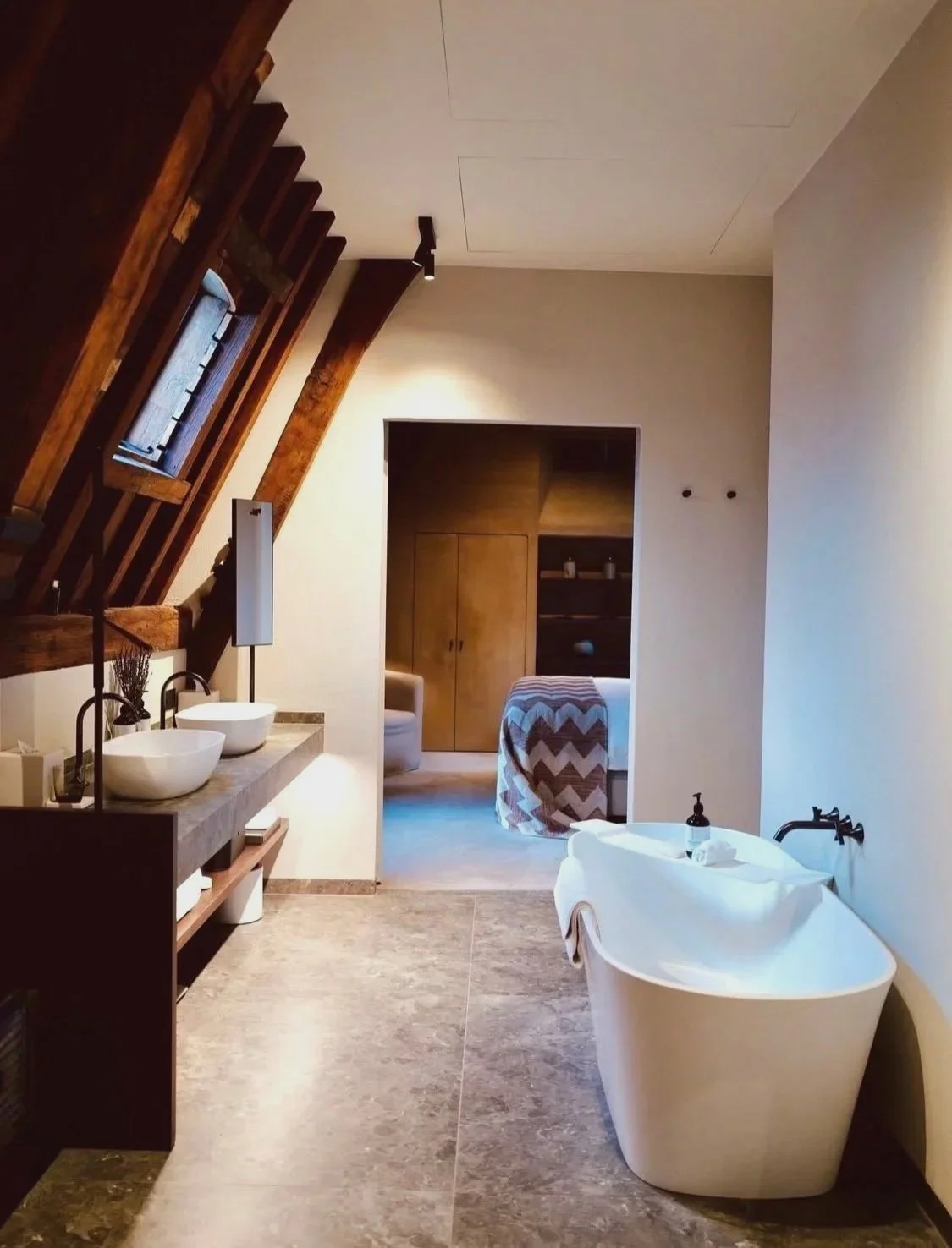 Modern bathroom with a white free-standing bathtub on the right, double sinks with vessel basins on a wooden vanity on the left, and a small window with wooden framing above. The room has beige tiled flooring, white walls, and a wooden ceiling. An open doorway leads to a bedroom with wooden furniture and a chevron-patterned blanket.