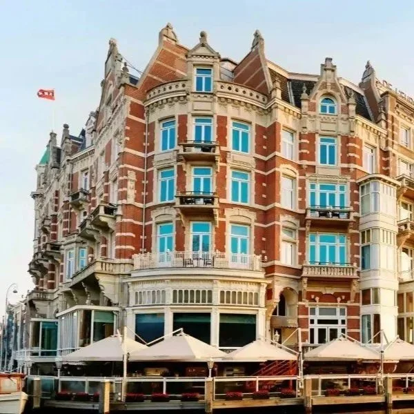 A historic multi-story red brick and cream-colored building with ornate architectural details, large windows, and balconies, situated along a waterfront with a pier and outdoor seating area with umbrellas.