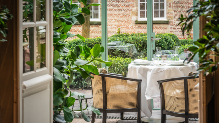 An outdoor dining table set for three people with white tablecloth, plates, glasses, and cutlery, seen through a glass door surrounded by greenery and a brick building in the background.