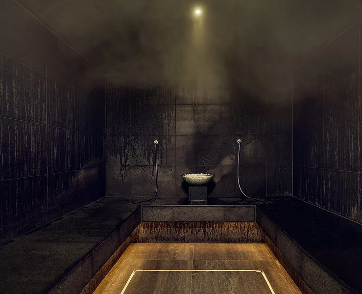 A dark, steam-filled traditional Japanese or Korean bathhouse room with black tiled walls, a small basin at the back, and steam rising from the basin.