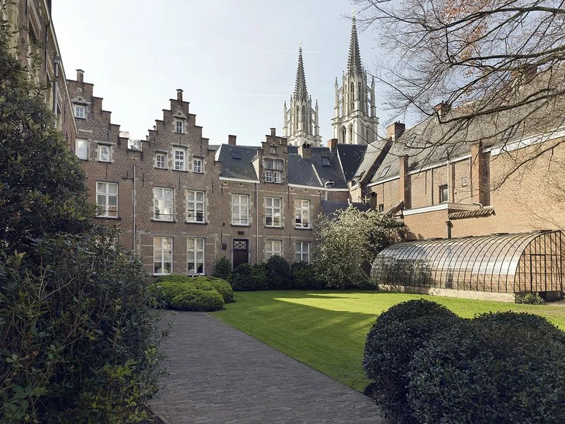 A courtyard with well-manicured grass, shrubs, a brick pathway, a greenhouse, surrounded by old brick buildings, with a church with twin steeples in the background.
