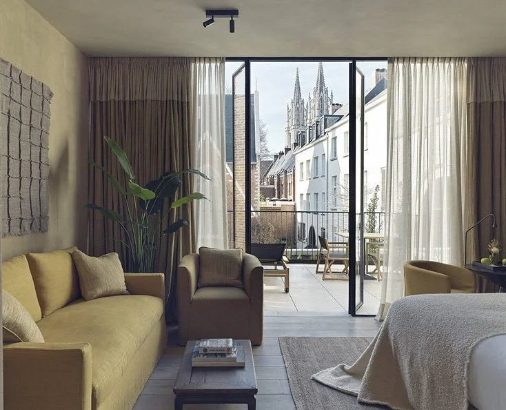 A living room with large glass sliding doors leading to a balcony, showing a view of historic church spires outside. The room is furnished with a yellow sofa, a beige armchair, a coffee table, and various plants and decor.