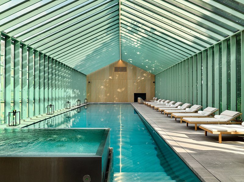 Indoor swimming pool with glass walls and ceiling, surrounded by white lounge chairs with wooden frames, natural light streaming in.