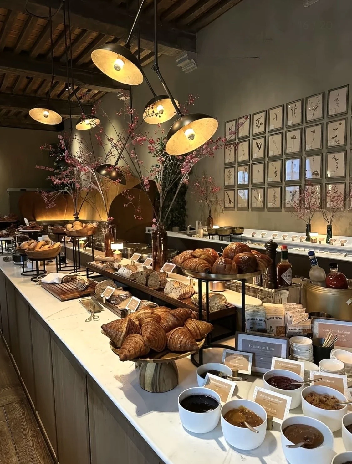 Dessert and bread buffet with croissants, various jams, and baked goods in a warmly lit, rustic-style cafe with framed art on the wall and decorative branches with pink blossoms.