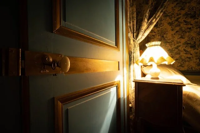 A partially open door leading into a dimly lit bedroom with a bedside table, a lit lamp, and a bed with floral bedding.