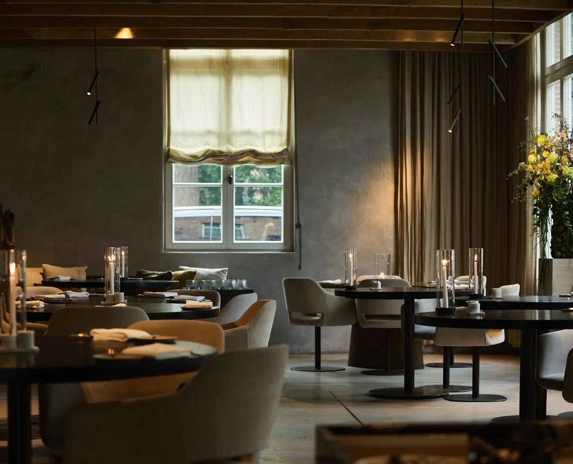 An elegantly set dining area inside a restaurant with round tables, beige chairs, tall candles, and a large window with curtains, illuminated by soft ambient lighting.
