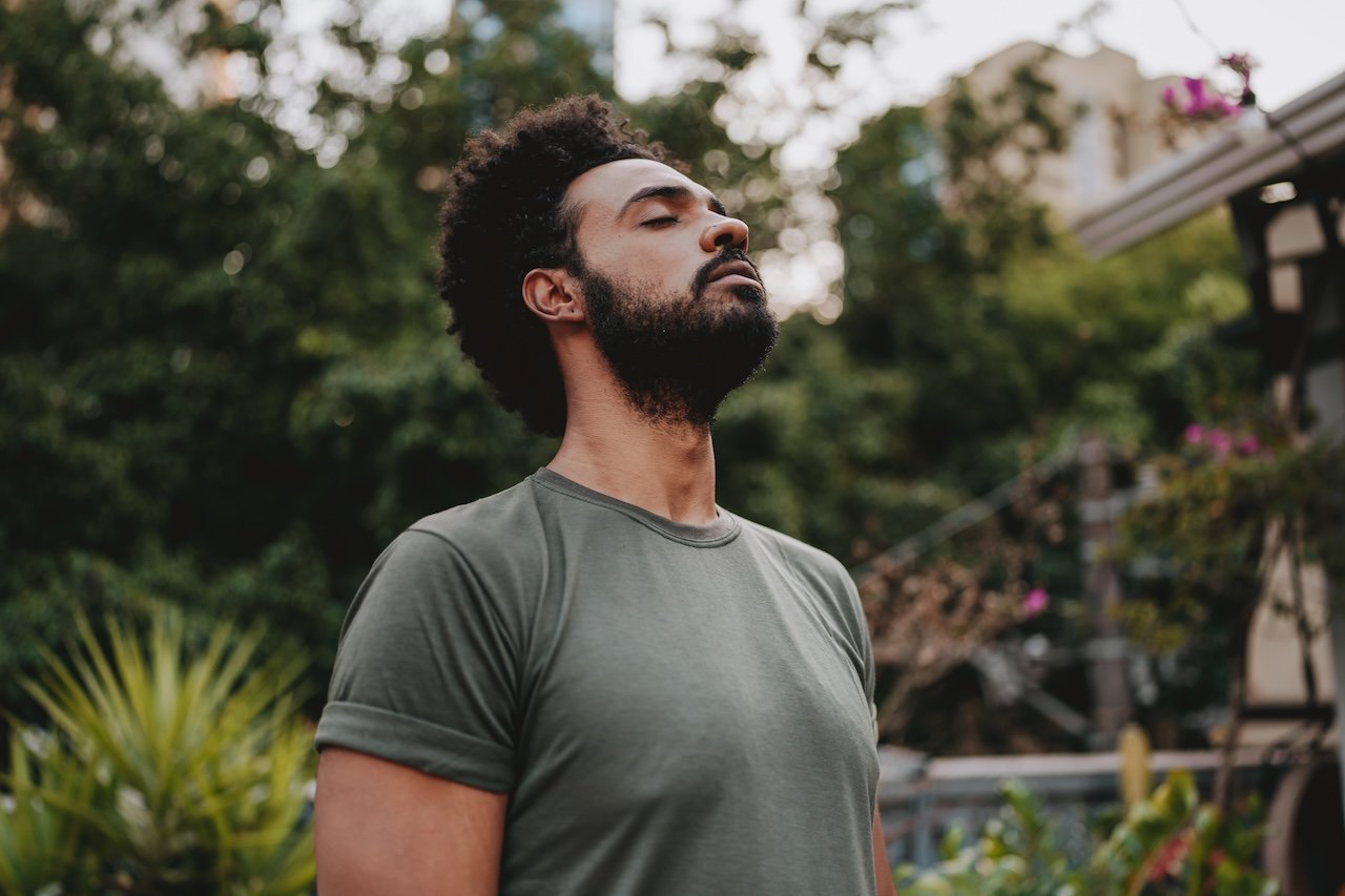A man with a beard and curly hair wearing a gray t-shirt standing outdoors with eyes closed and a peaceful expression.