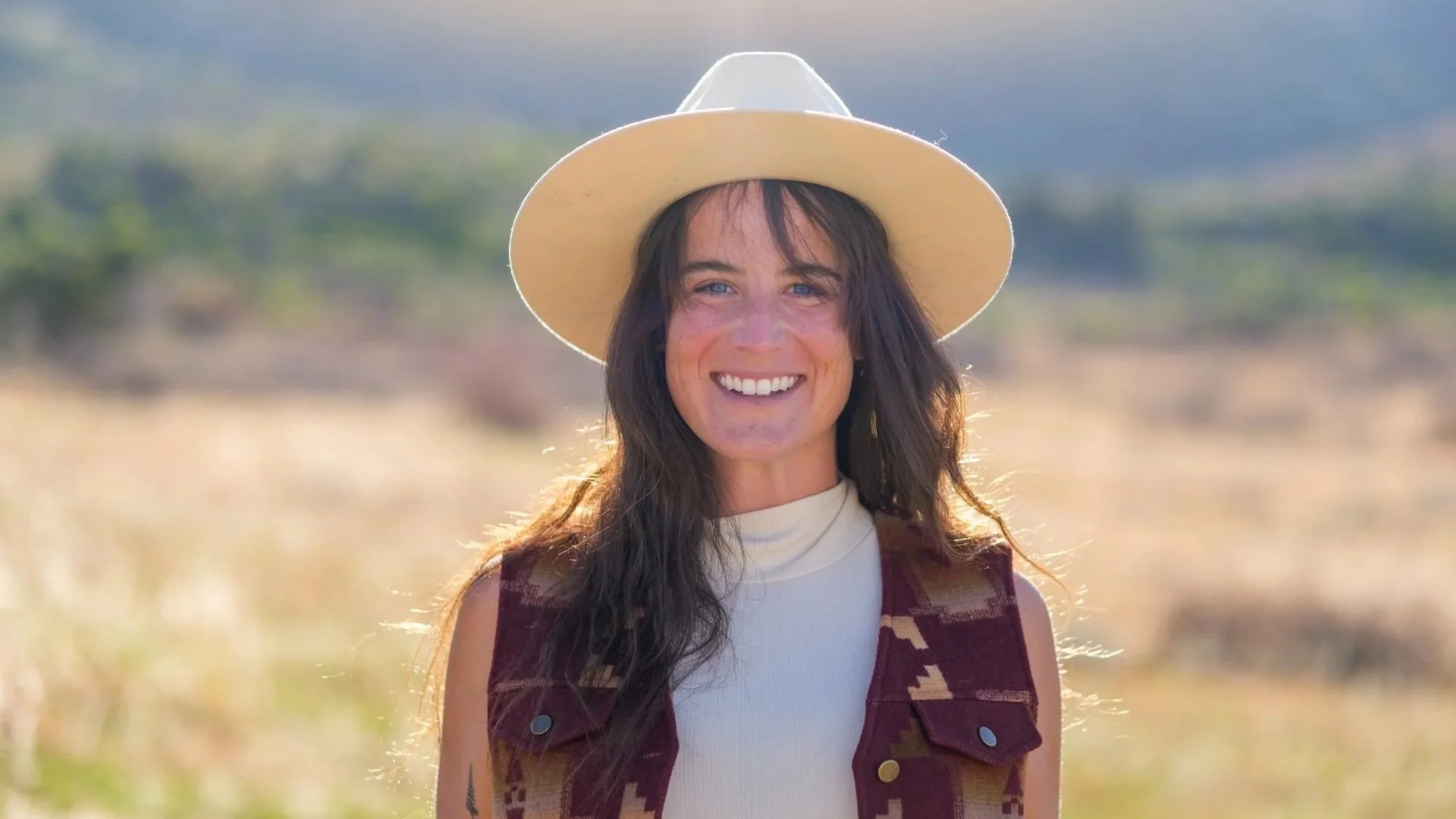 A woman with long dark hair and blue eyes smiling outdoors during daytime. She is wearing a wide-brimmed hat, a white sleeveless top, and a brown patterned vest. The background is blurred with natural scenery.