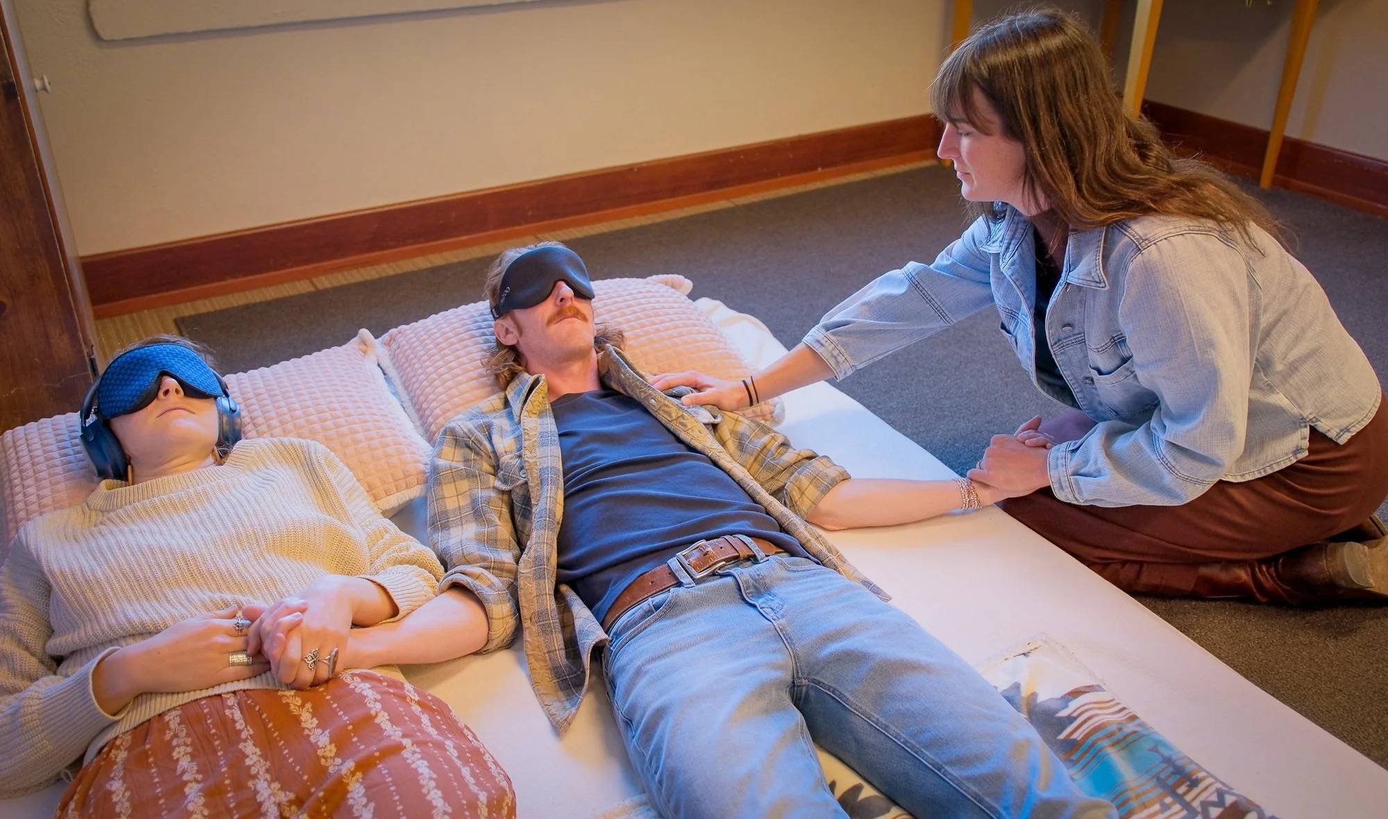 A woman kneeling next to a bed, holding the hand of a man lying on the bed with an eye mask over his eyes, while another woman lying next to them appears relaxed.
