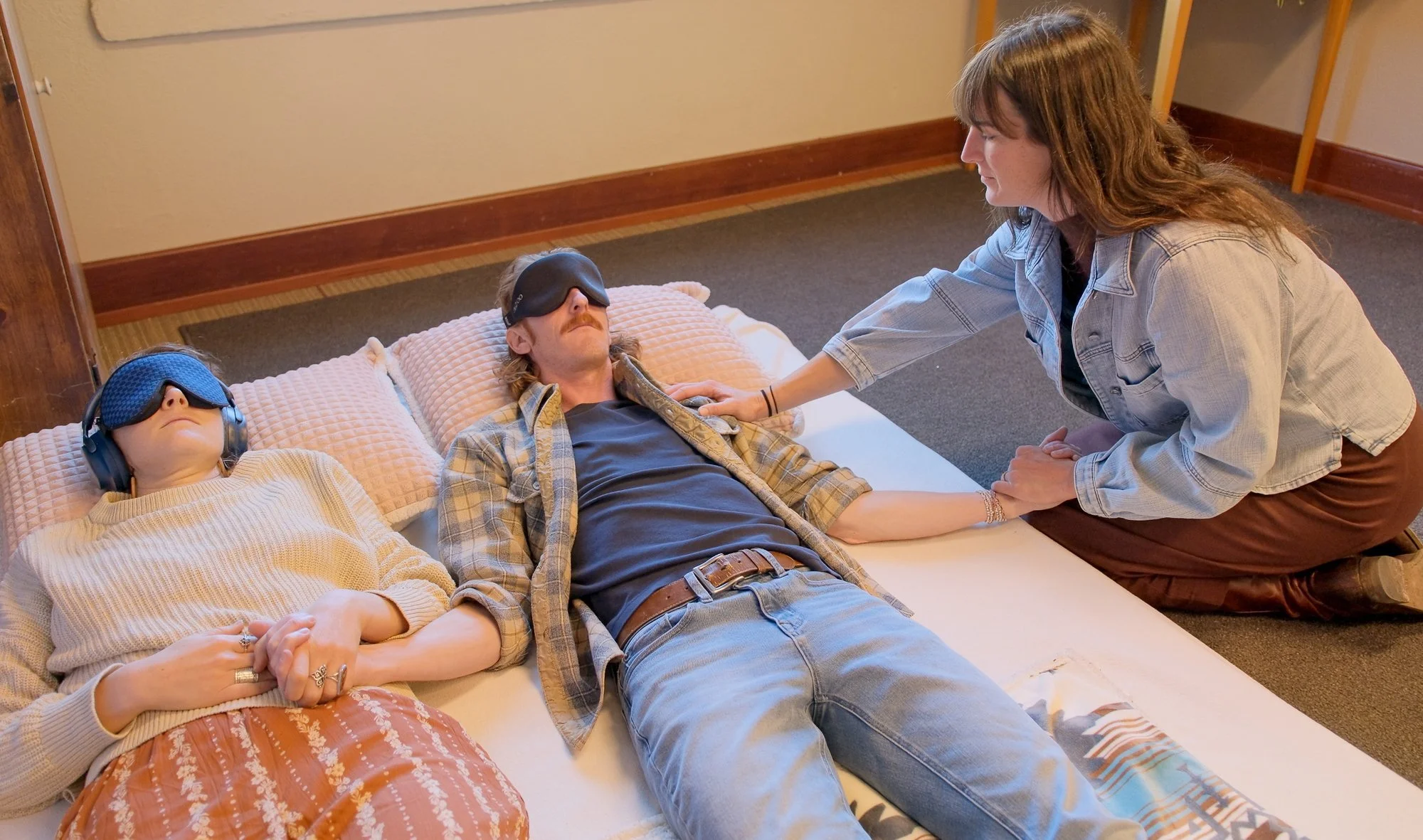 A woman is kneeling beside two people lying on a bed, one man and one woman, all with blindfolds. The woman on the bed is holding hands with the person beside her, and the woman kneeling is touching the shoulder of the man.