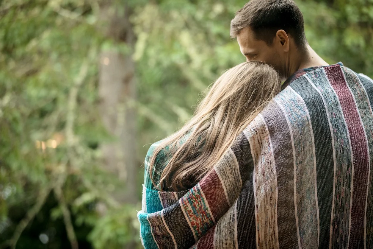 A man and woman embracing outdoors with the woman resting her head on the man's shoulder, wrapped in a striped blanket, in a green wooded area.