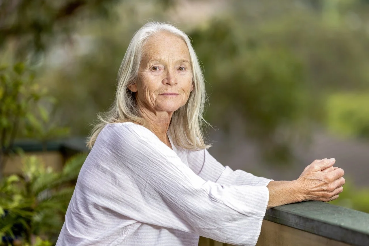 An elderly woman with long gray hair, wearing a white blouse, sitting outdoors on a wooden railing with greenery in the background.