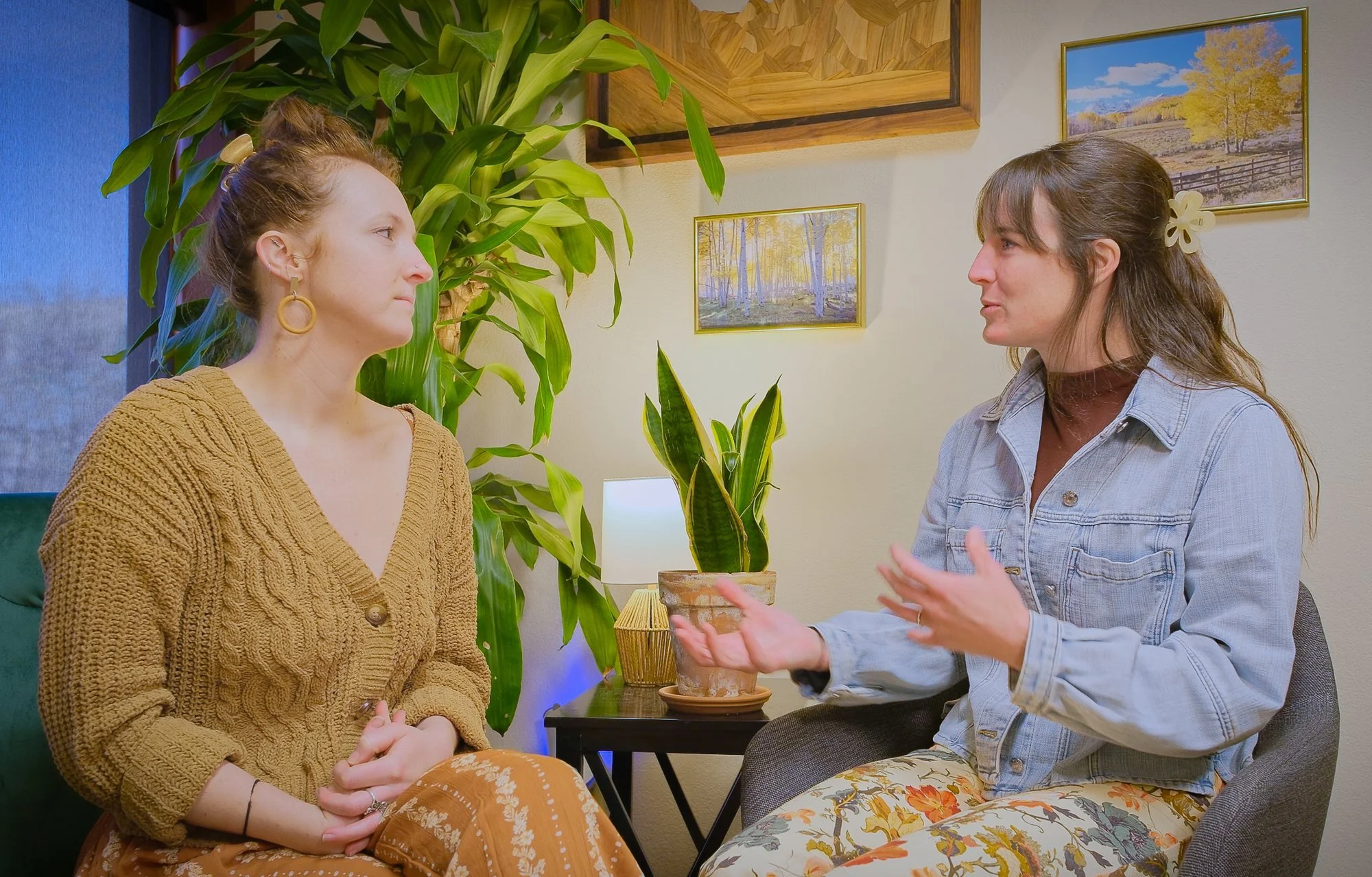 Two women are sitting and having a conversation indoors. One woman, on the left, has curly hair tied up with a hair clip, wearing a mustard yellow knitted cardigan and patterned brown pants. The other woman, on the right, has brown hair tied back with a flower hair clip, wearing a denim jacket and floral pants. They are positioned near a table with potted plants and framed landscape art on the wall behind them. The conversation appears to be engaged and expressive.