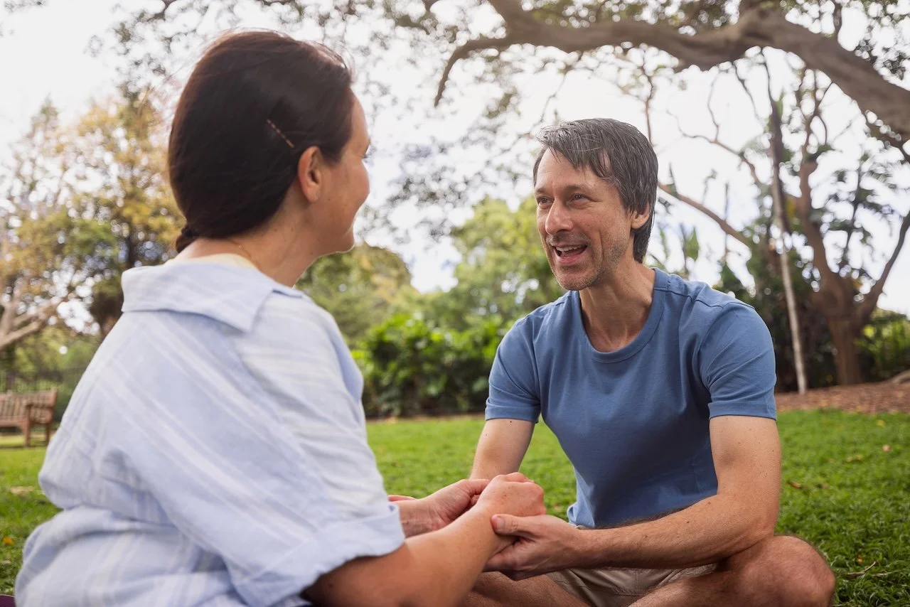 A man and woman hold hands and talk happily outdoors in a park with trees and grass.