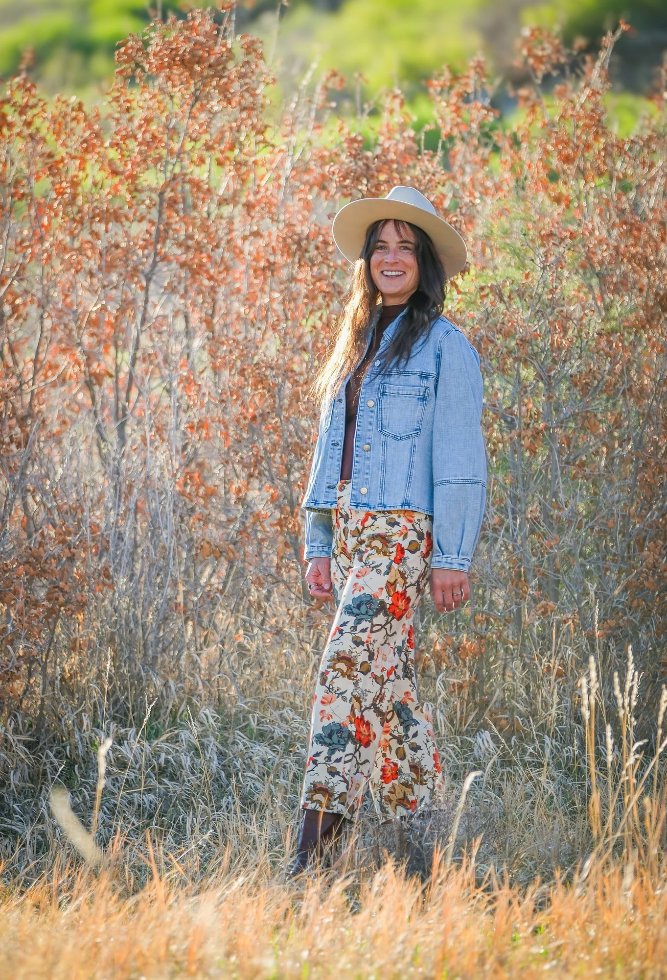 A woman with long dark hair, smiling, wearing a wide-brimmed hat, denim jacket, and floral patterned pants, standing in a field of dried grass with orange and brown bushes in the background.