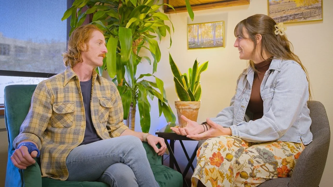 A woman and man engaging in a conversation in a cozy room with yellow walls, framed pictures, and large green plants. The woman is smiling with open hands, and the man is listening attentively.