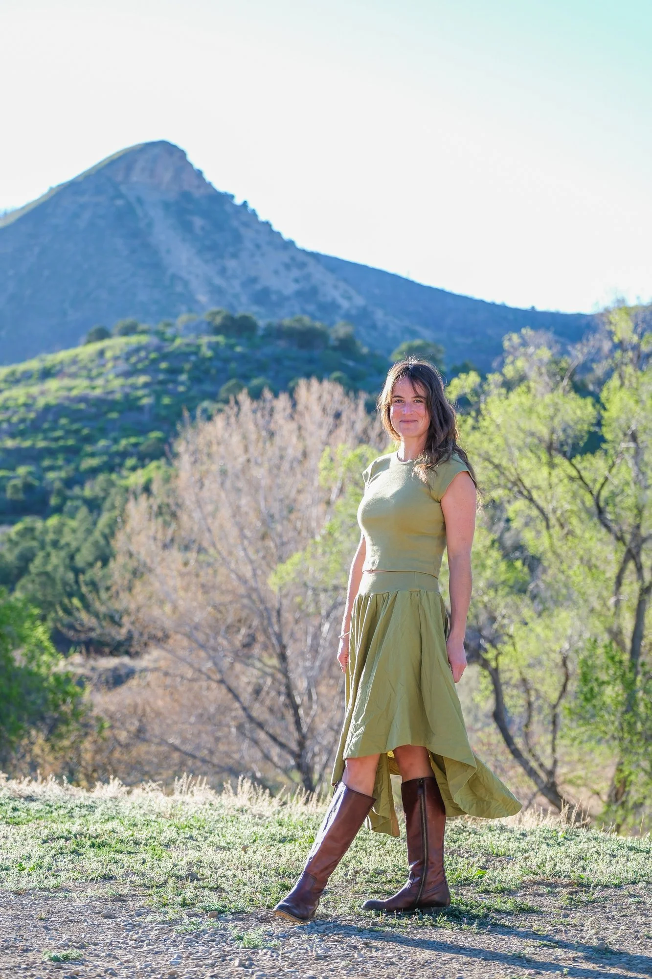 A woman standing outdoors on a sunny day with a mountain in the background, wearing a light green dress and tall brown boots.